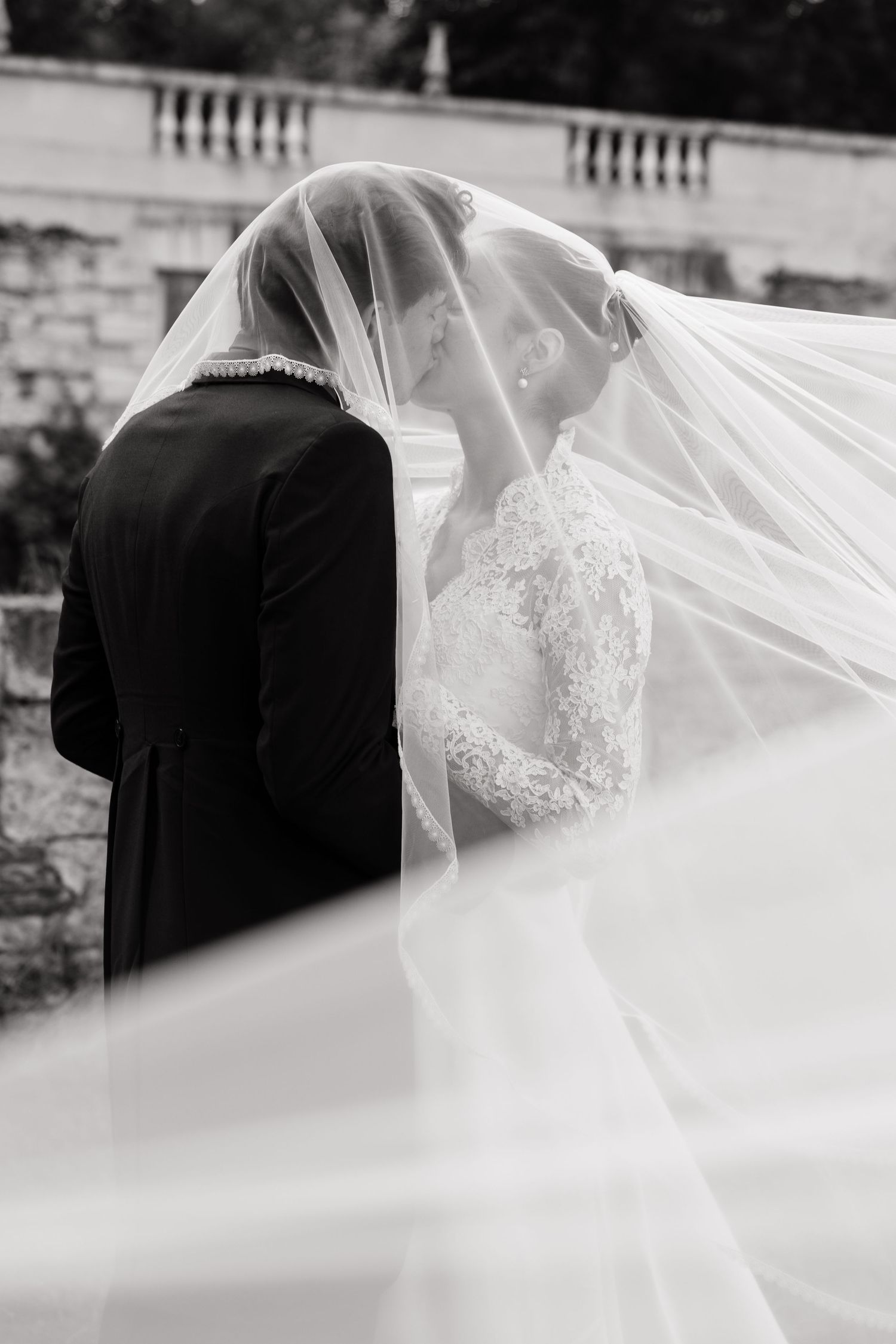 Black and white photo of bride and groom sharing a kiss under a flowing veil at an elegant outdoor wedding.