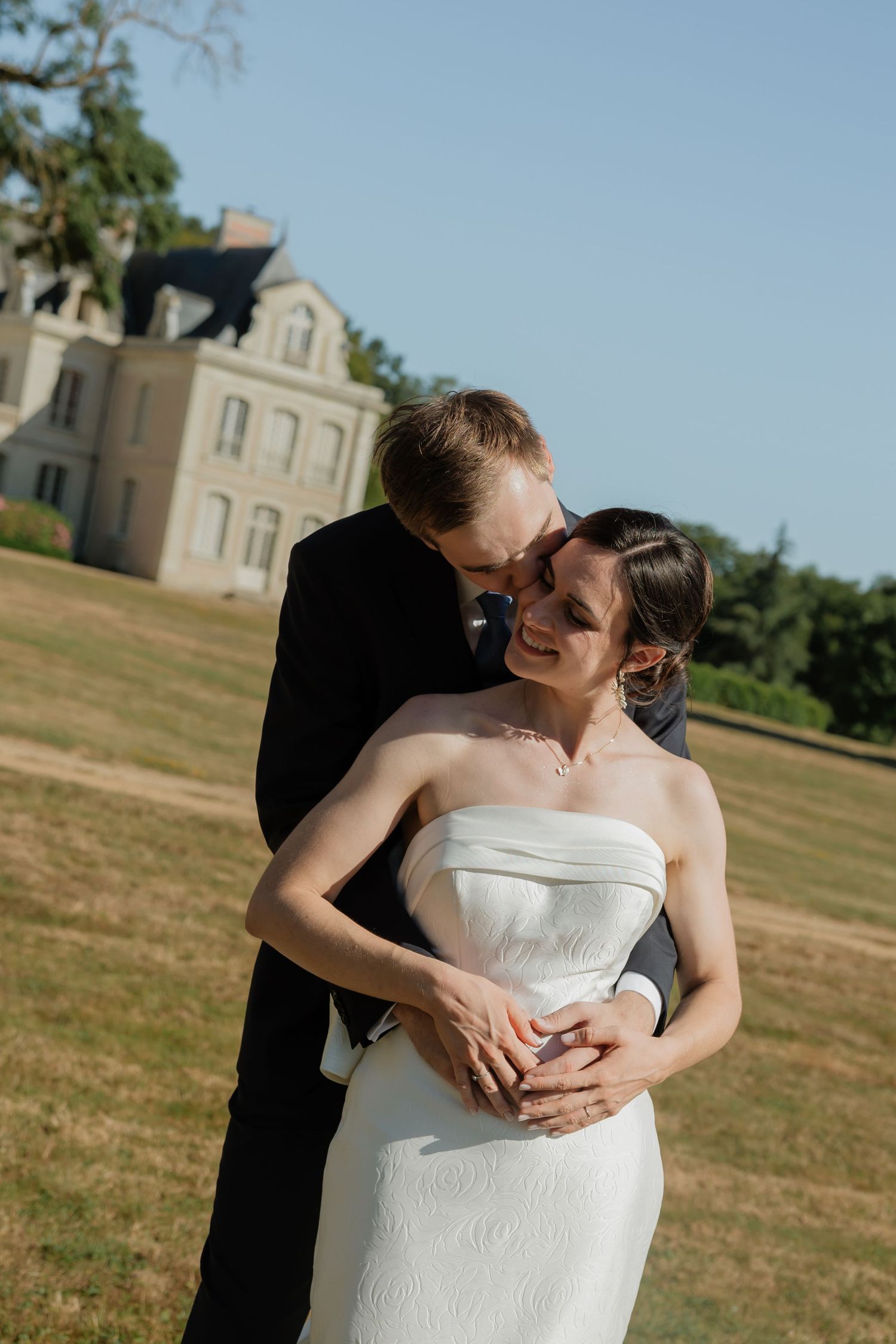 A groom in a black suit embraces his smiling bride in a white strapless gown outdoors near a elegant estate.