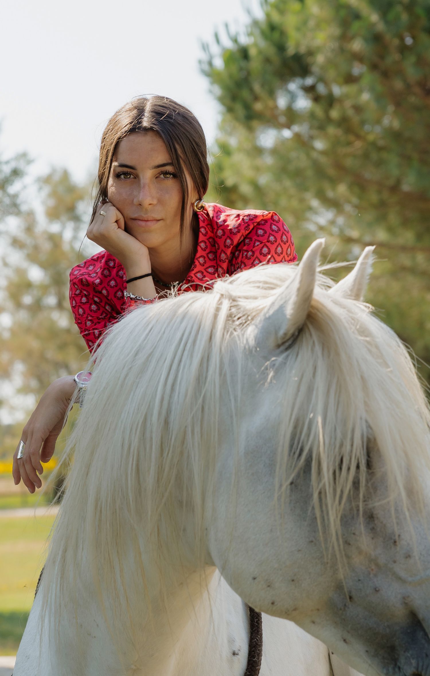 Young woman in red patterned top leaning on a white horse outdoors with trees in the background.