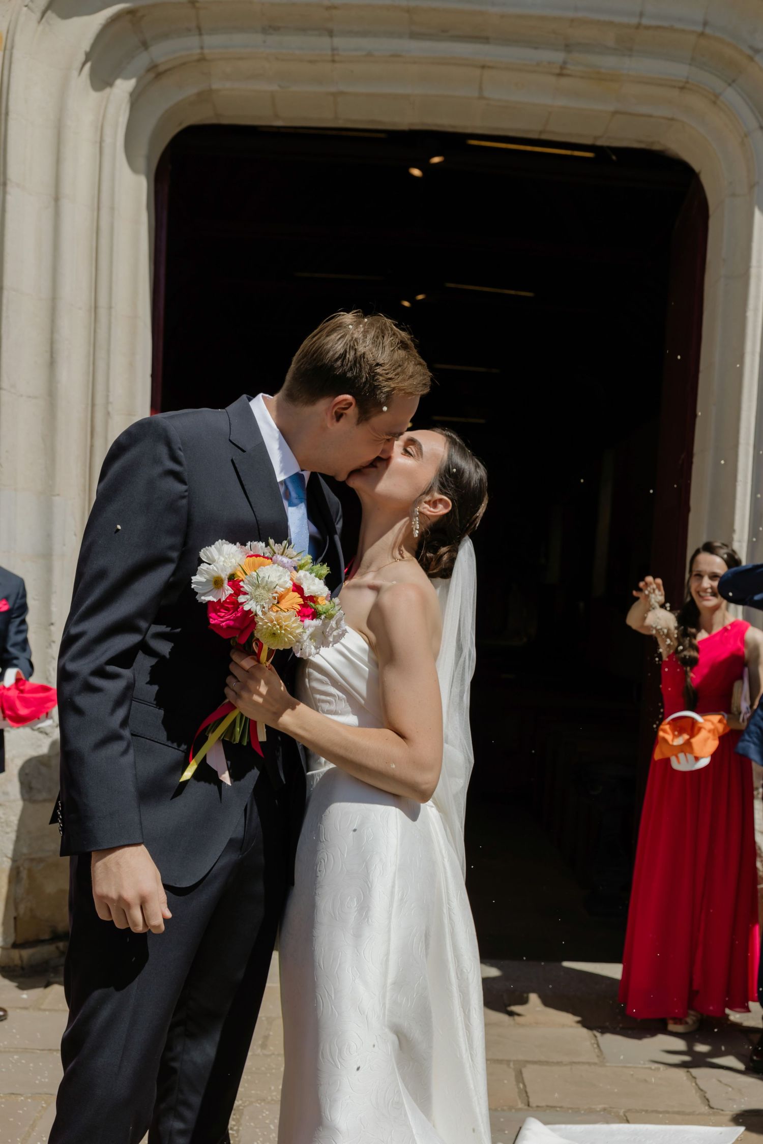 Bride and groom share first kiss outside church doors, bride holding colorful bouquet, guests celebrating in background.