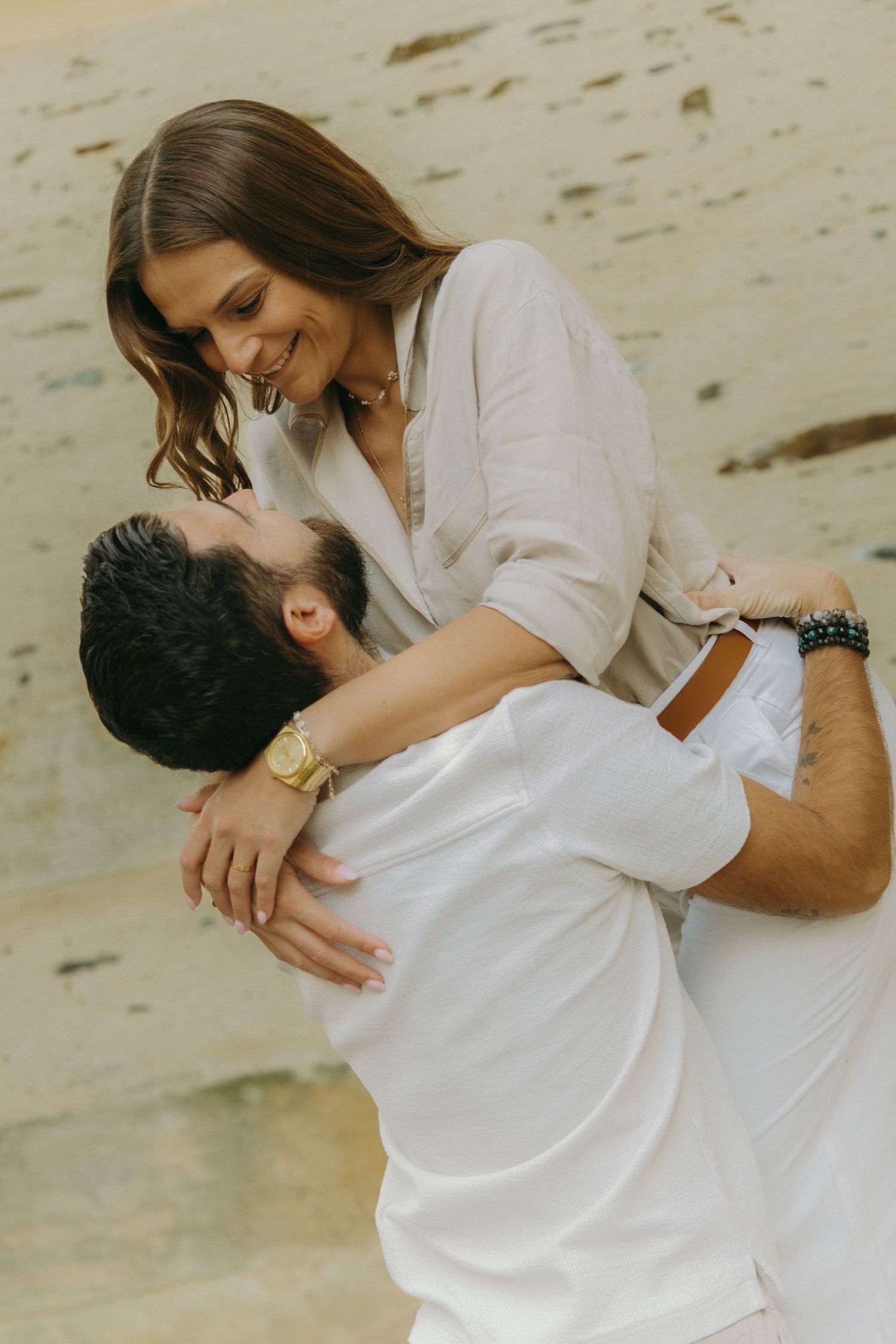 A couple dressed in white embraces tenderly on a sandy beach.