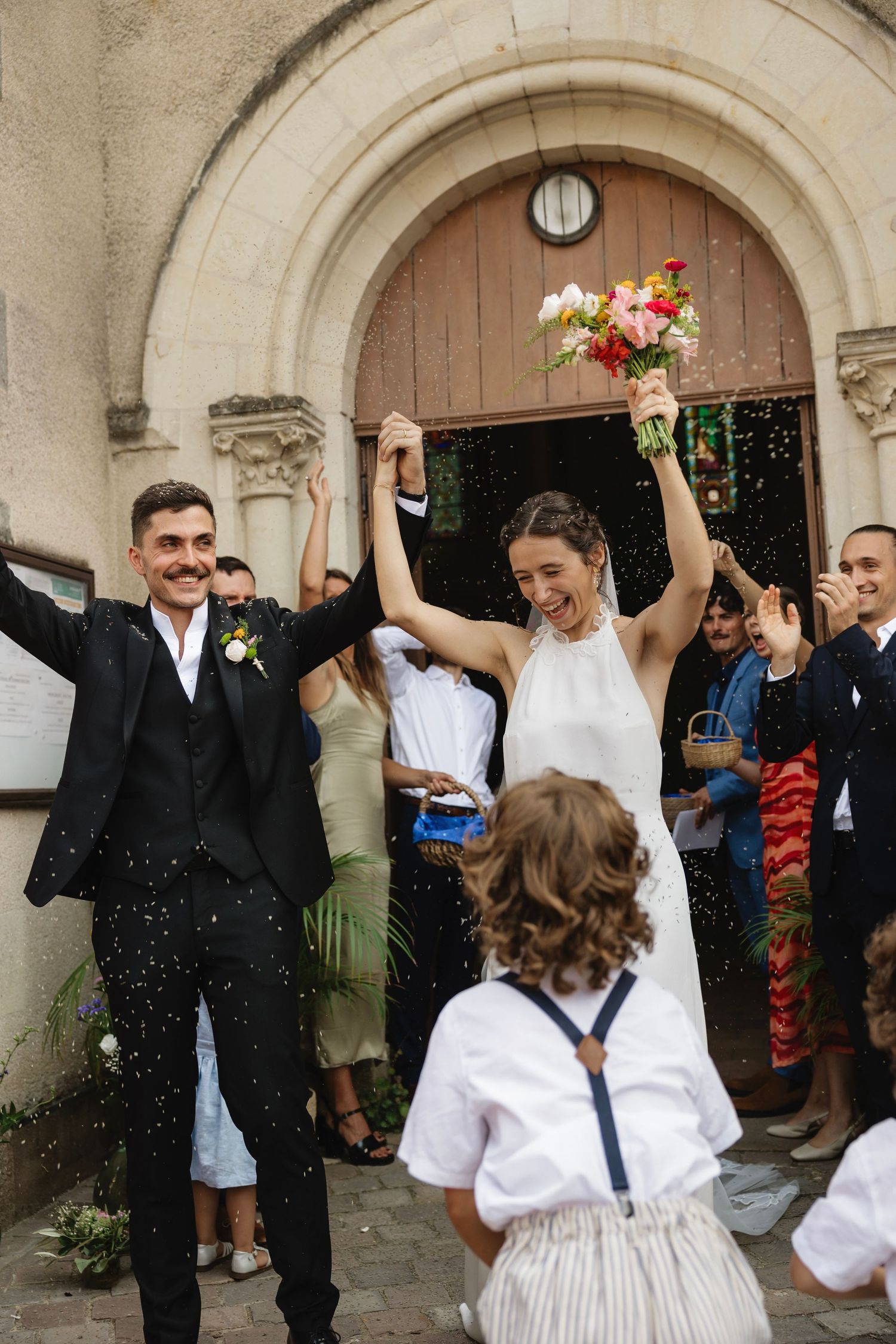 Joyful newlyweds celebrate outside church, bride holds colorful bouquet aloft as guests shower them with confetti.