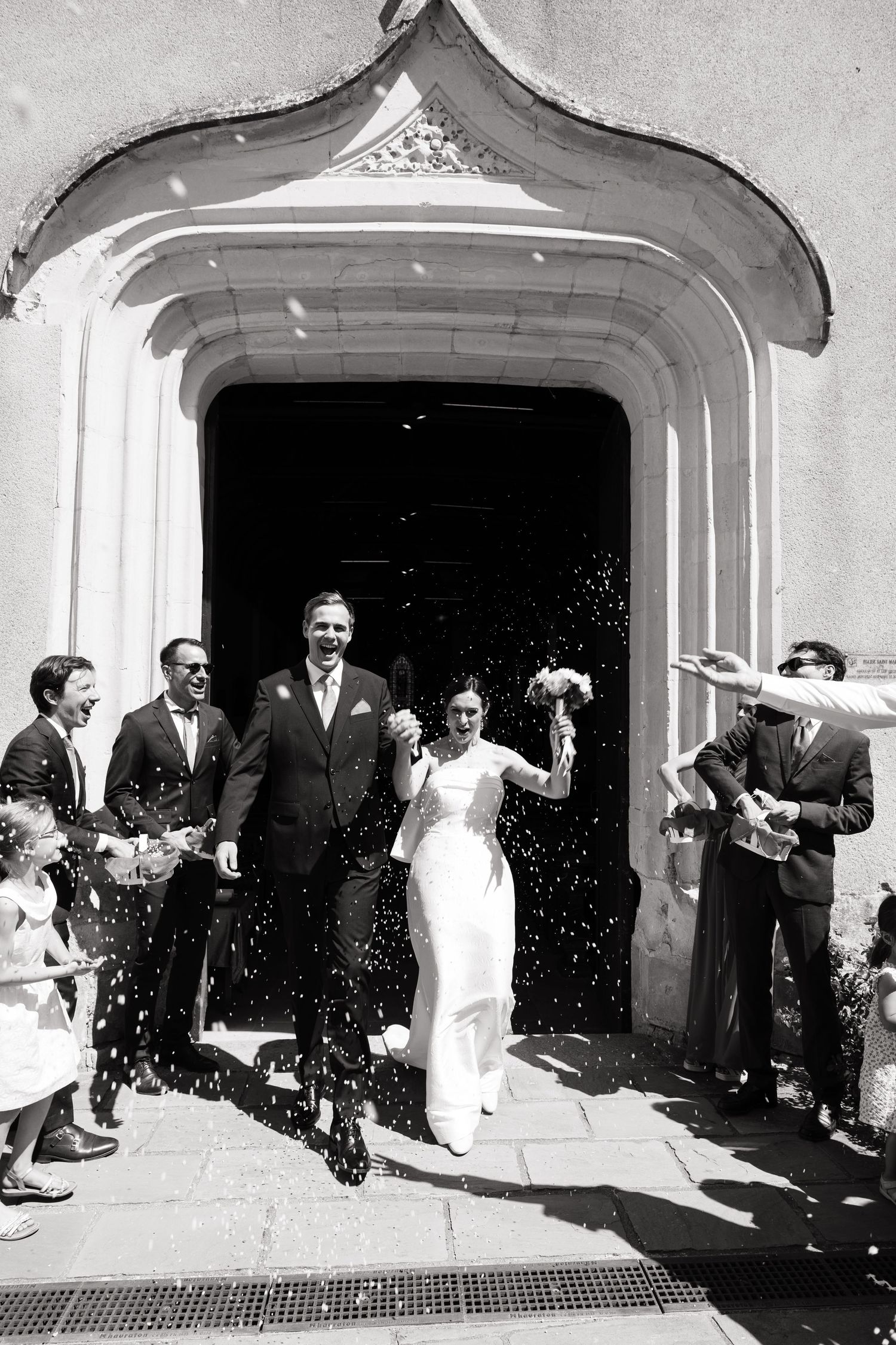 Black and white photo of a newlywed couple exiting a church as guests throw confetti in celebration.