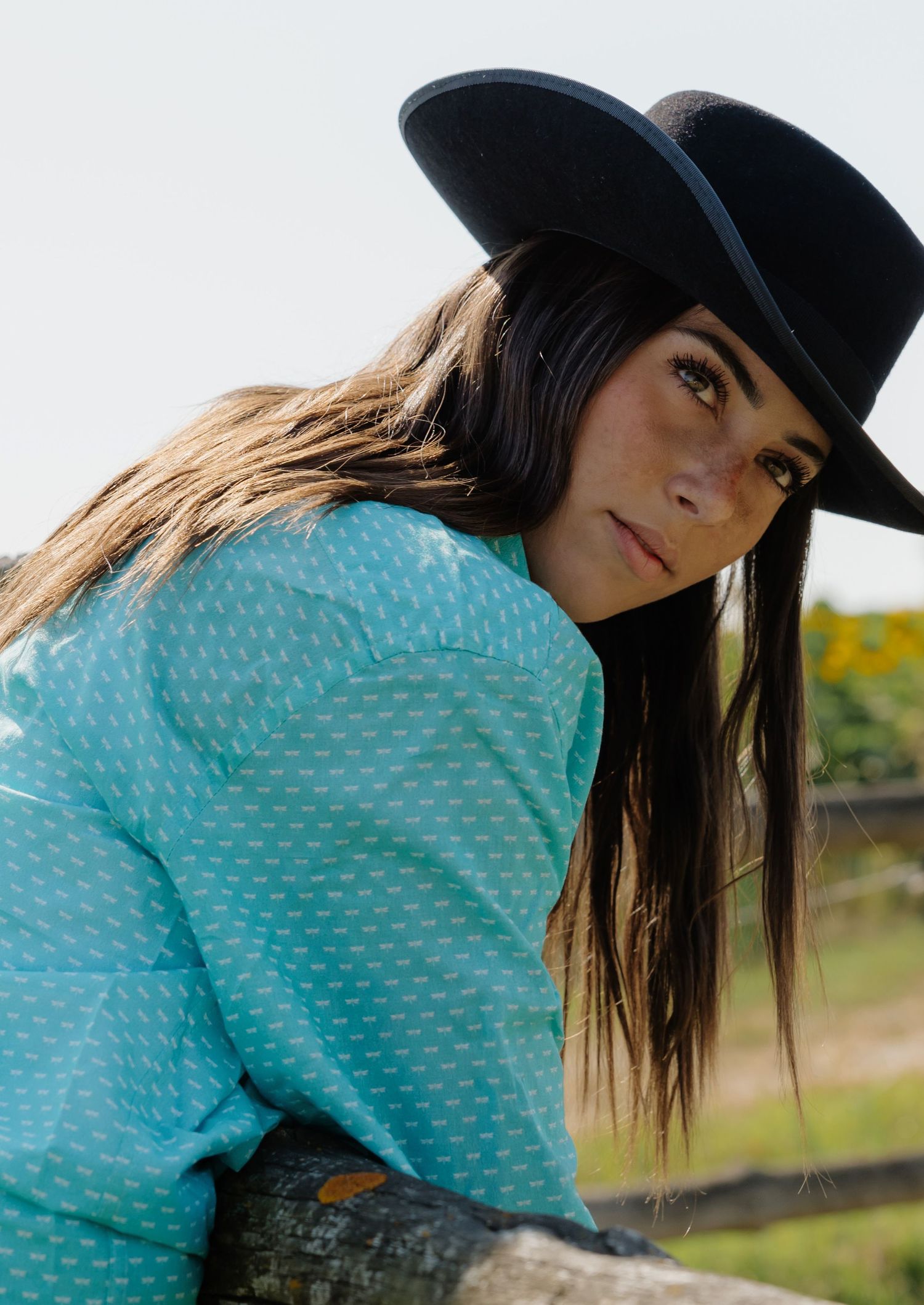 Young woman in black wide-brim hat and turquoise patterned shirt leaning on a wooden fence outdoors.