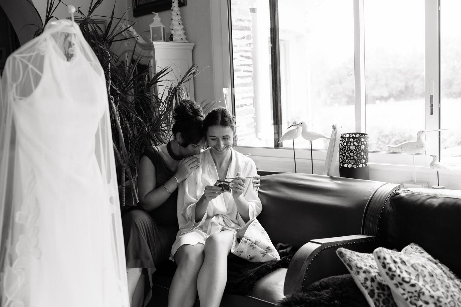 Bride in white robe sits on leather sofa checking phone with bridesmaid, wedding dress hanging nearby, black and white photo.