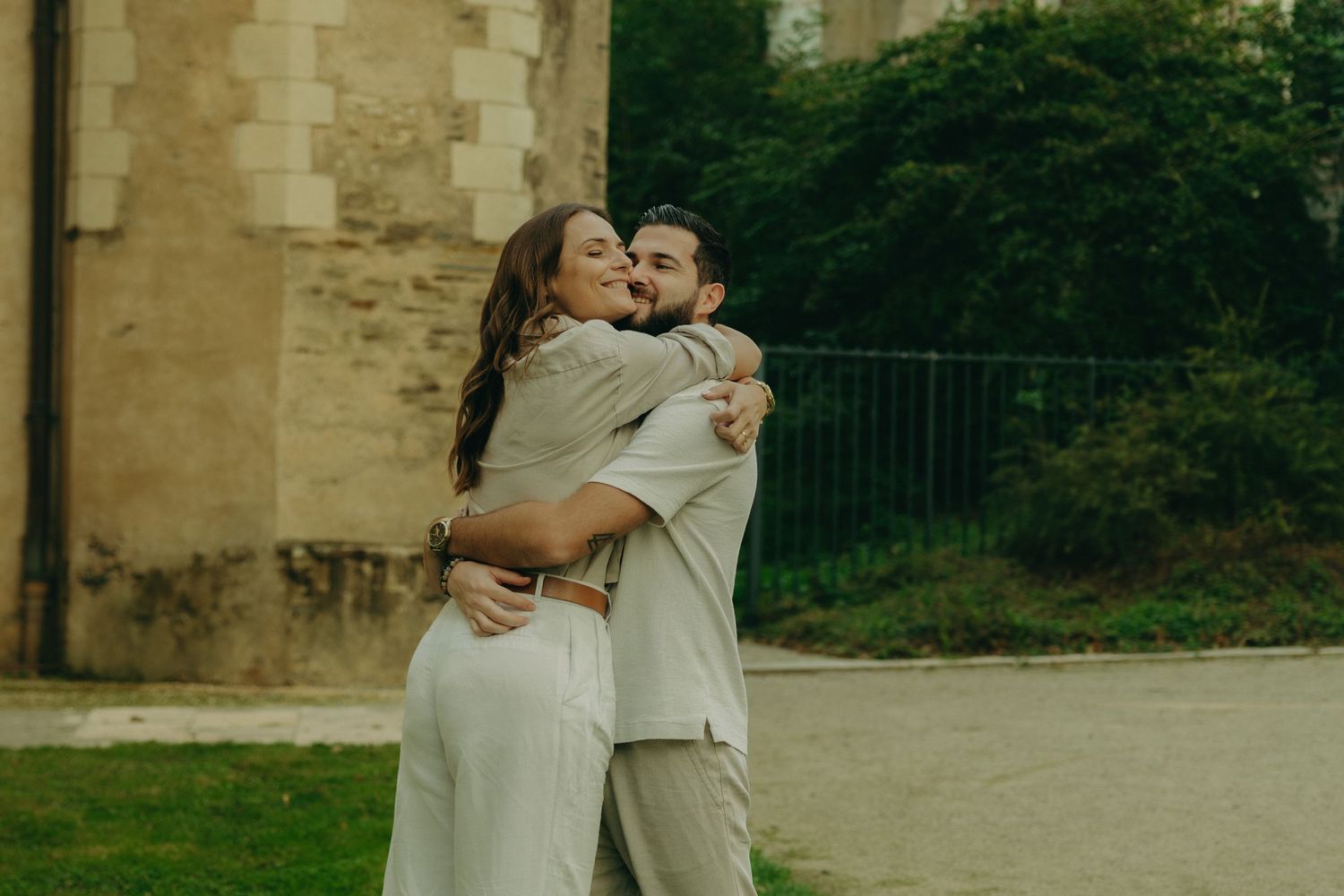 Two people in white outfits embrace and dance together in front of a stone building.