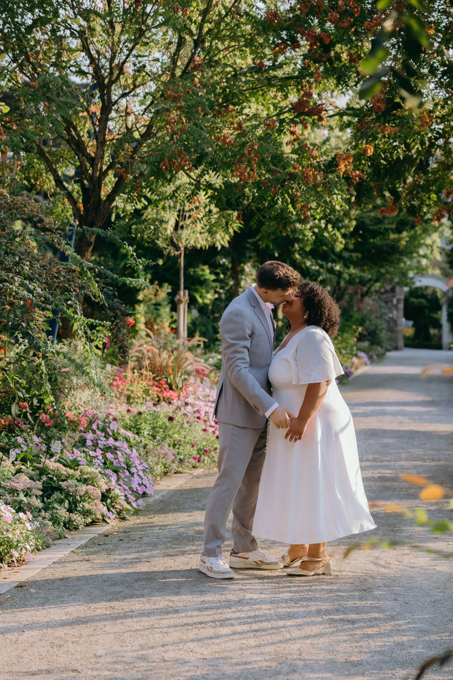 Un couple en tenue de mariage s’enlace sur un chemin de jardin bordé de fleurs colorées. Arboretum d'angers