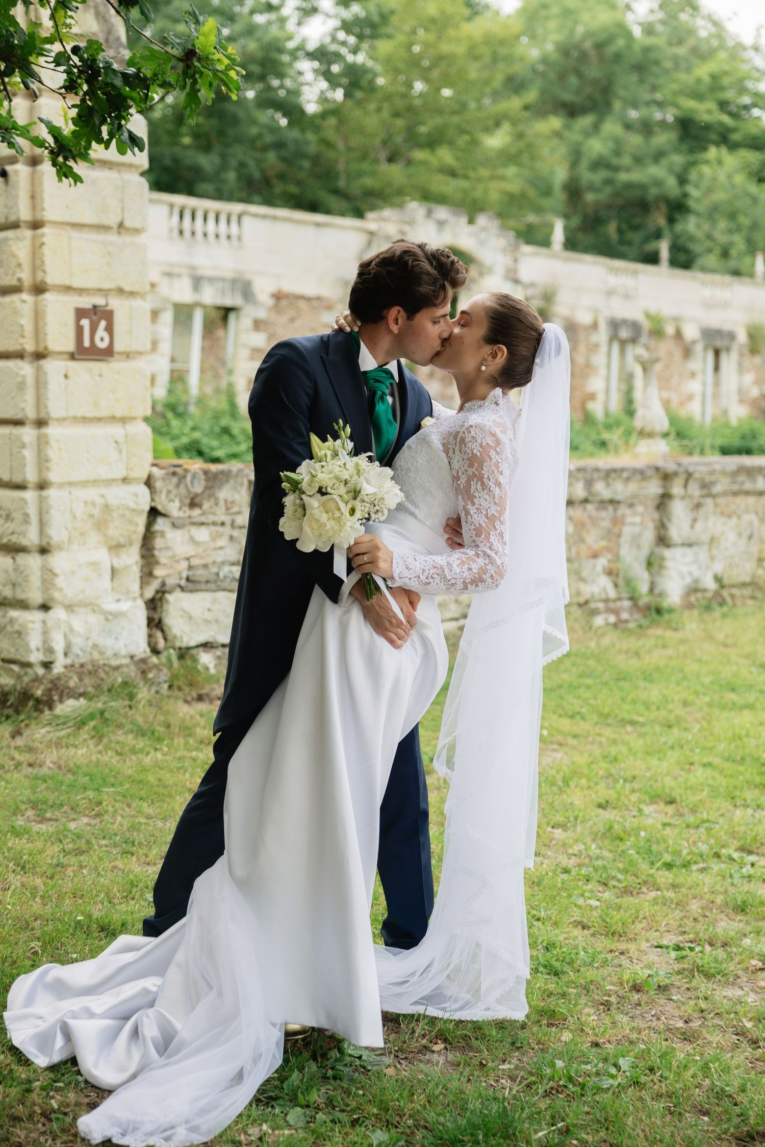 Bride and groom share a romantic kiss outdoors, bride in lace gown with veil holding white bouquet near stone building.
