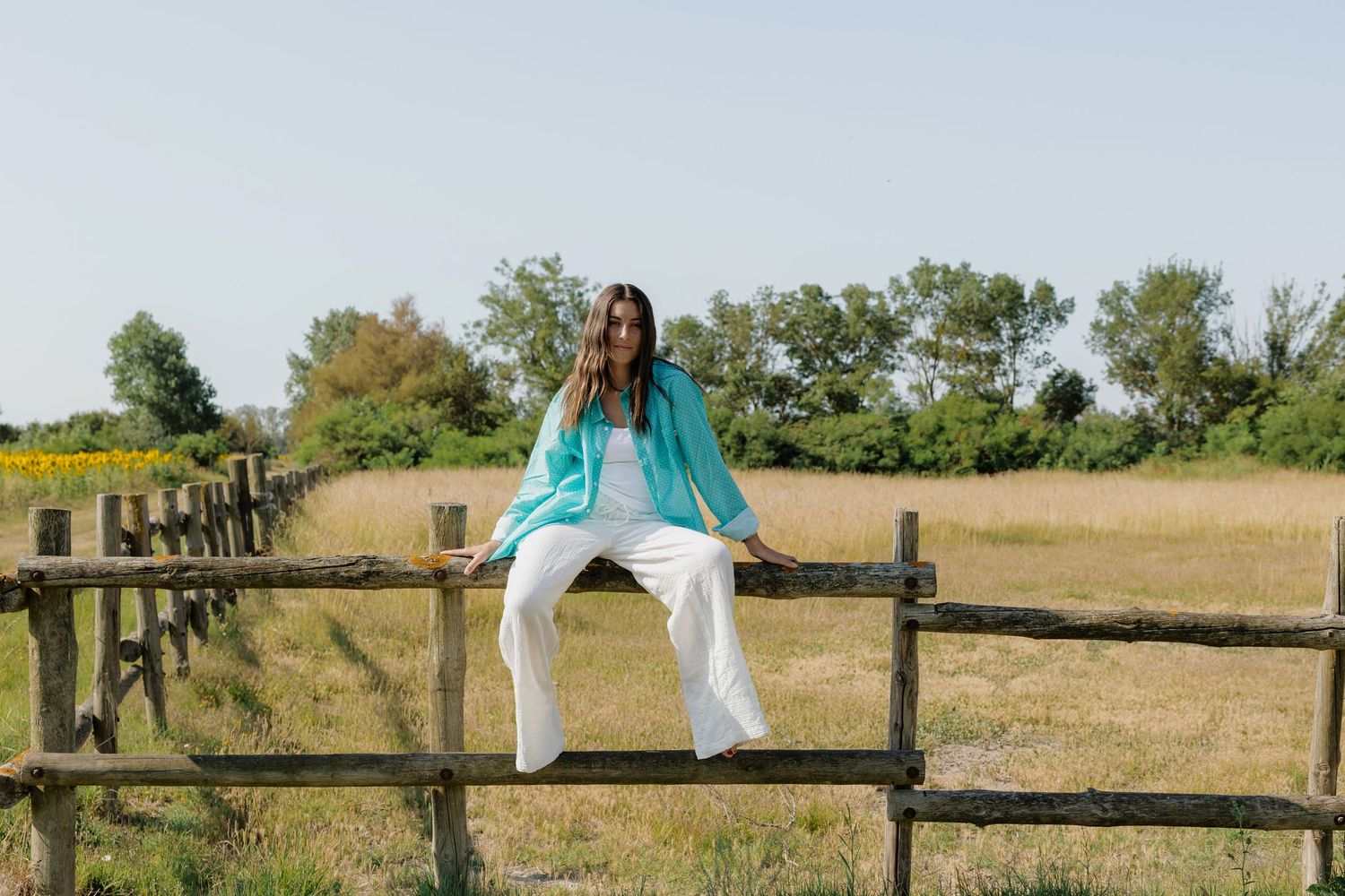 Woman in turquoise top and white pants sitting on a rustic wooden fence in a sunny rural field.