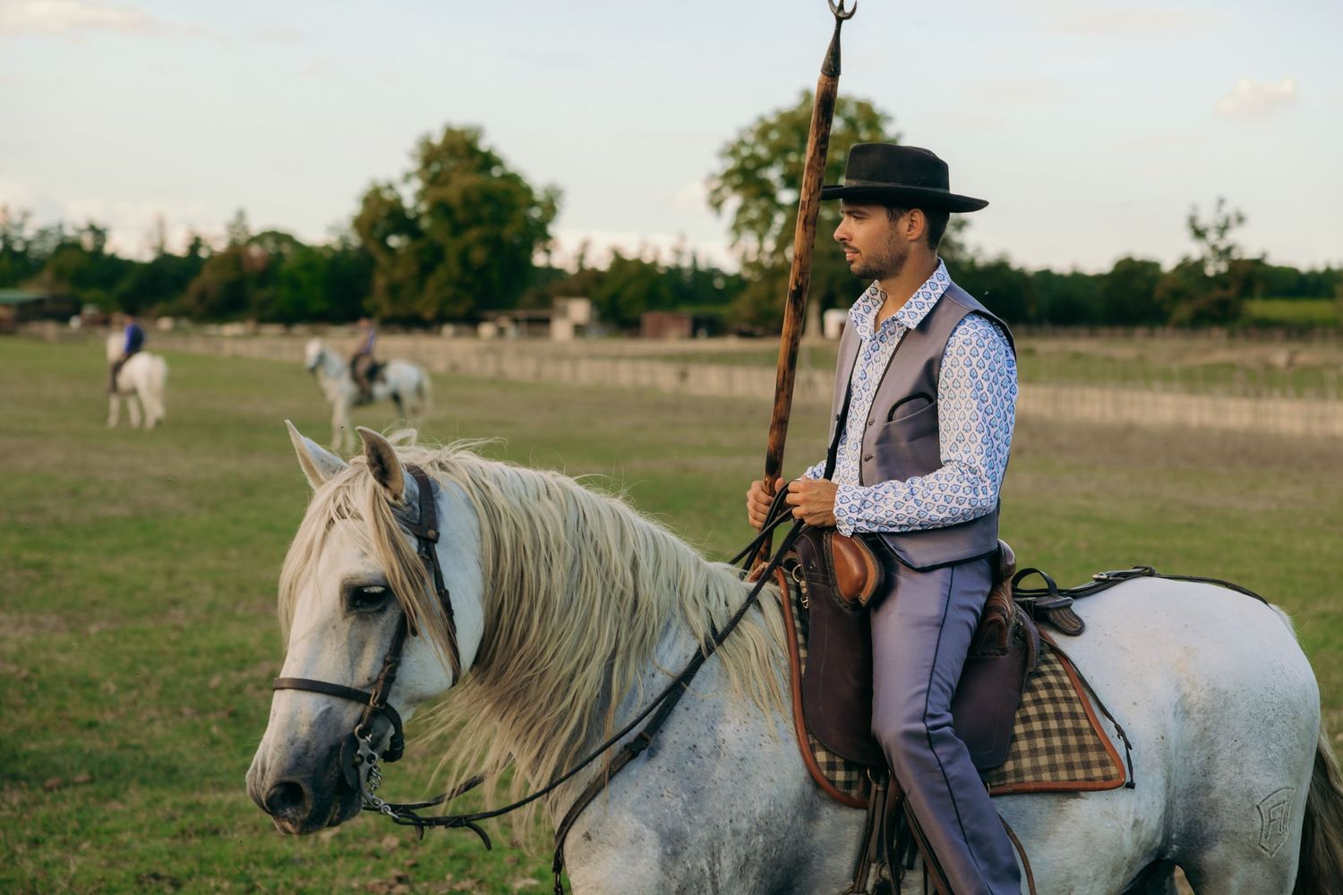 Riders on horseback move through a pastoral field with wooden fencing.