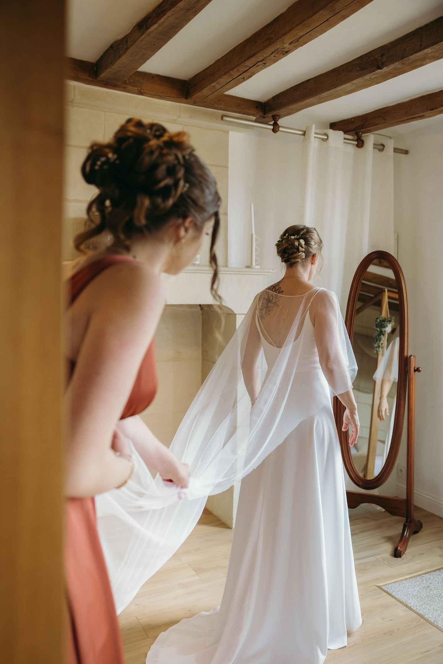 Photographie d’une mariée portant une robe blanche fluide style cape, se tenant devant un miroir ovale vintage.