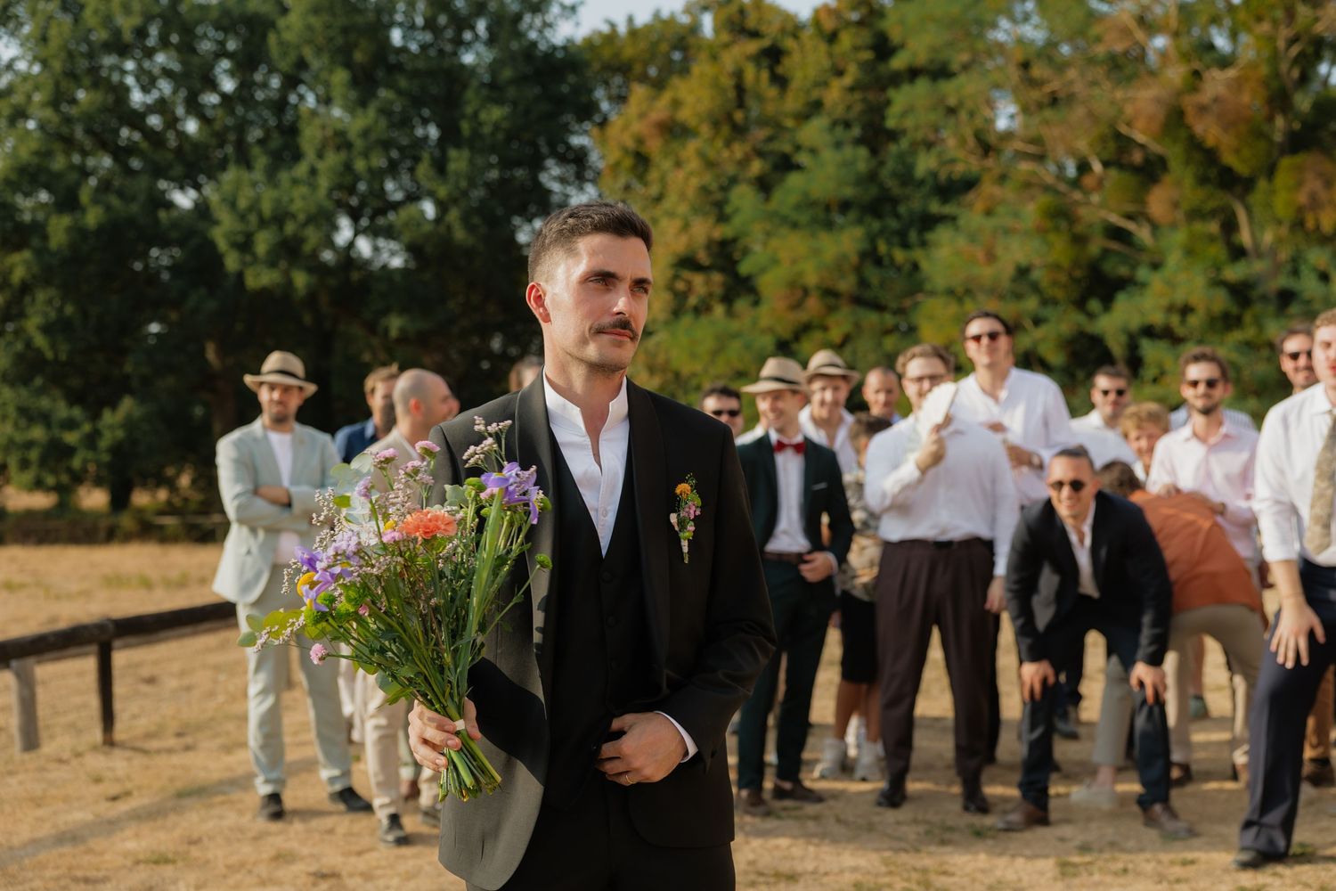 Groom in black suit holds colorful bouquet outdoors, with wedding guests gathered in background on sunny day.