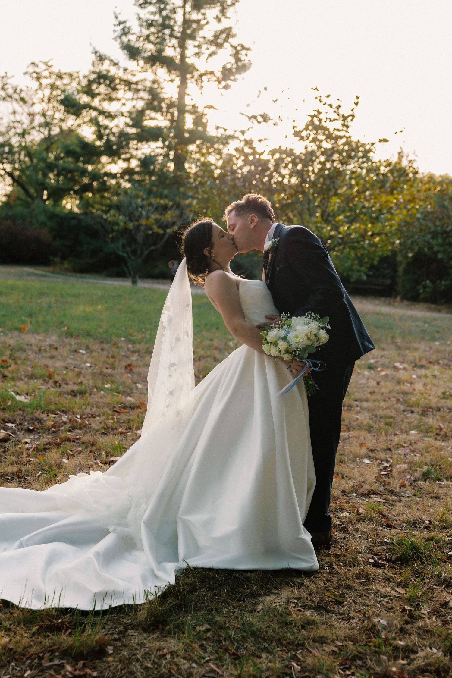 Bride and groom share a romantic kiss outdoors at golden hour, with bride holding white bouquet and wearing a flowing gown with veil.