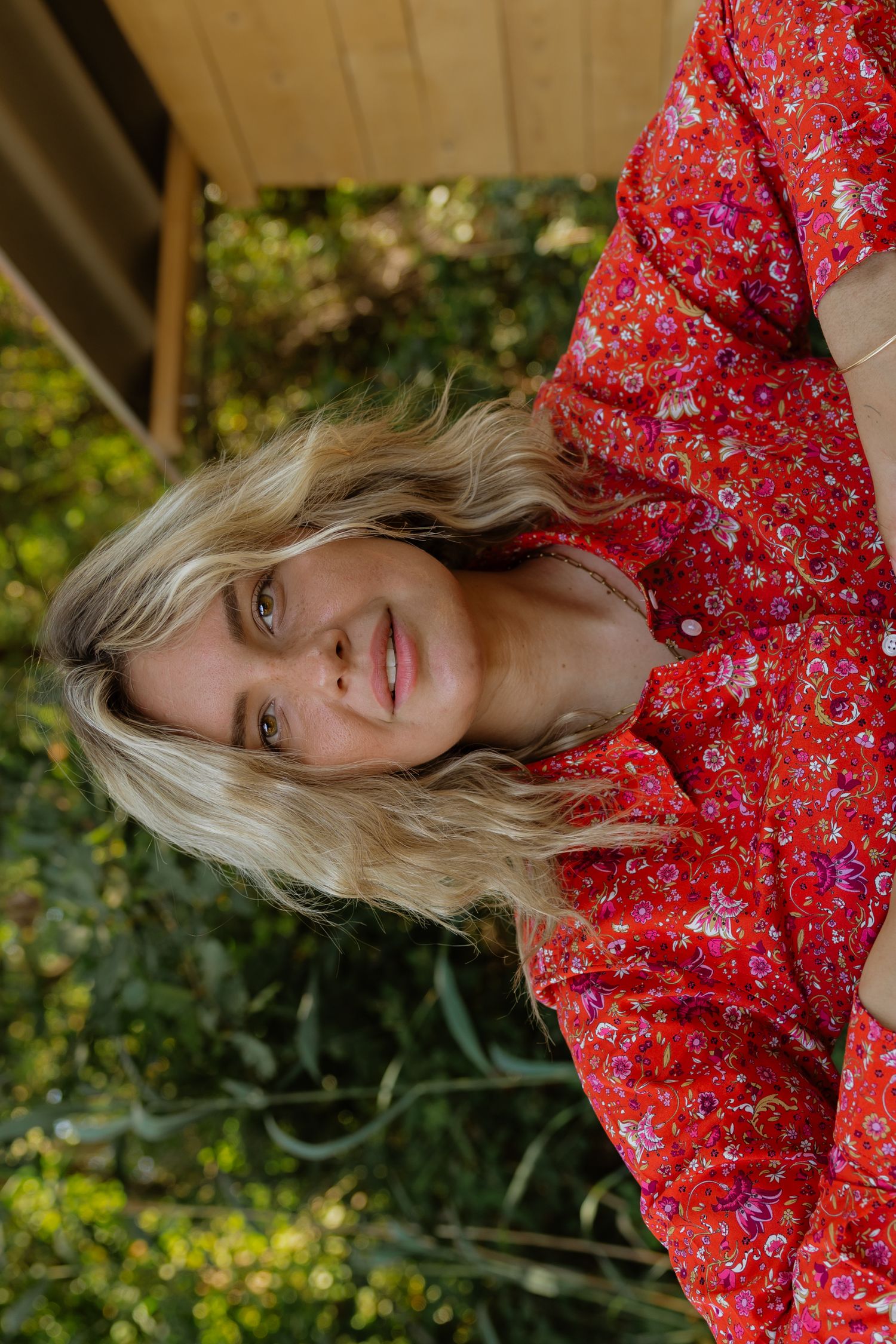 Blonde woman in red floral dress smiling outdoors surrounded by lush green garden foliage.