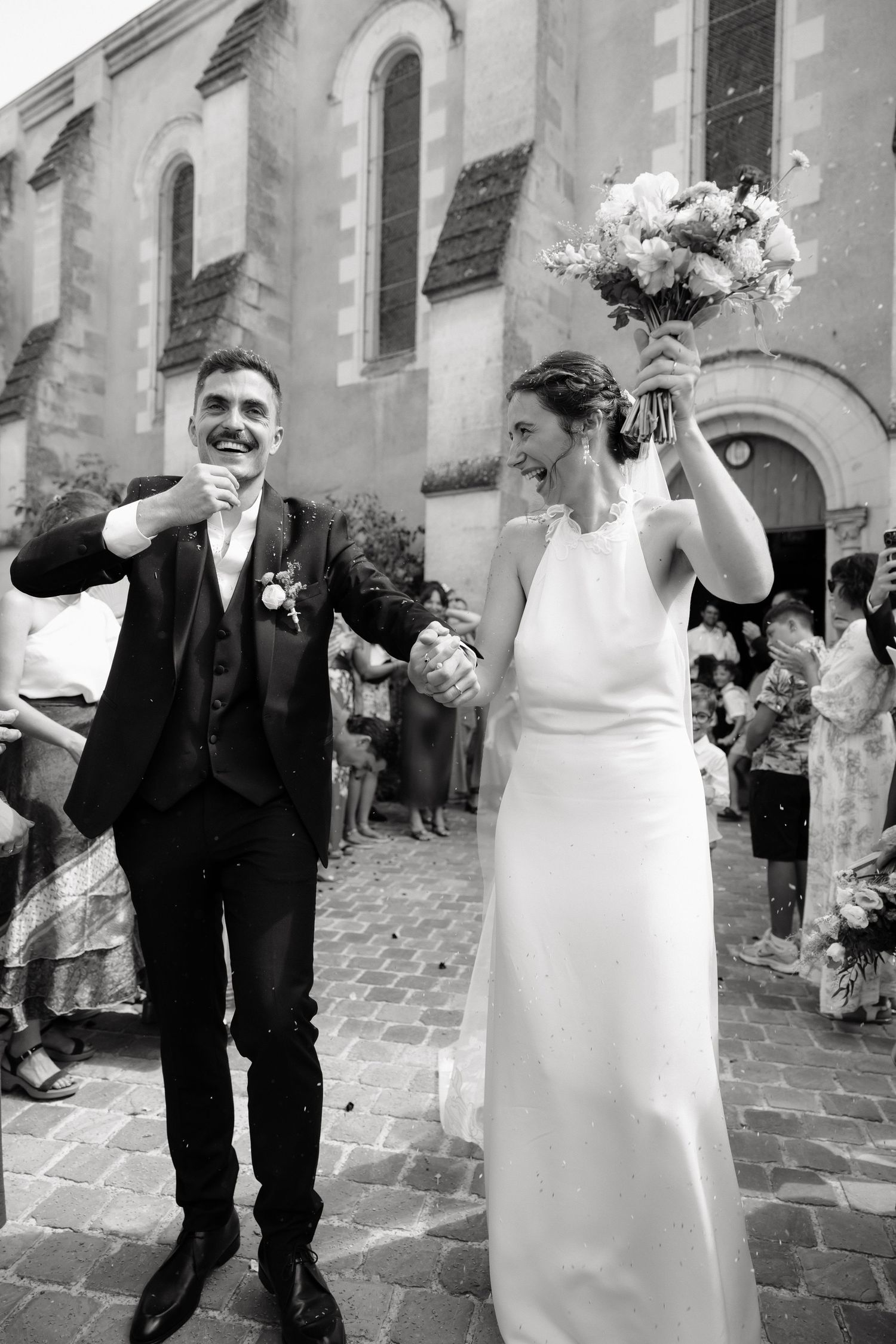 Black and white photo of joyful newlyweds exiting church, bride raising bouquet, guests celebrating in background.