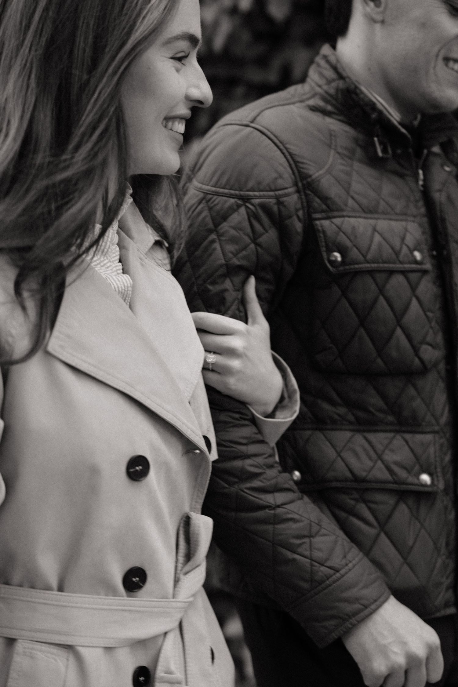 Black and white photo of a smiling woman in a trench coat walking with a man in a quilted jacket.