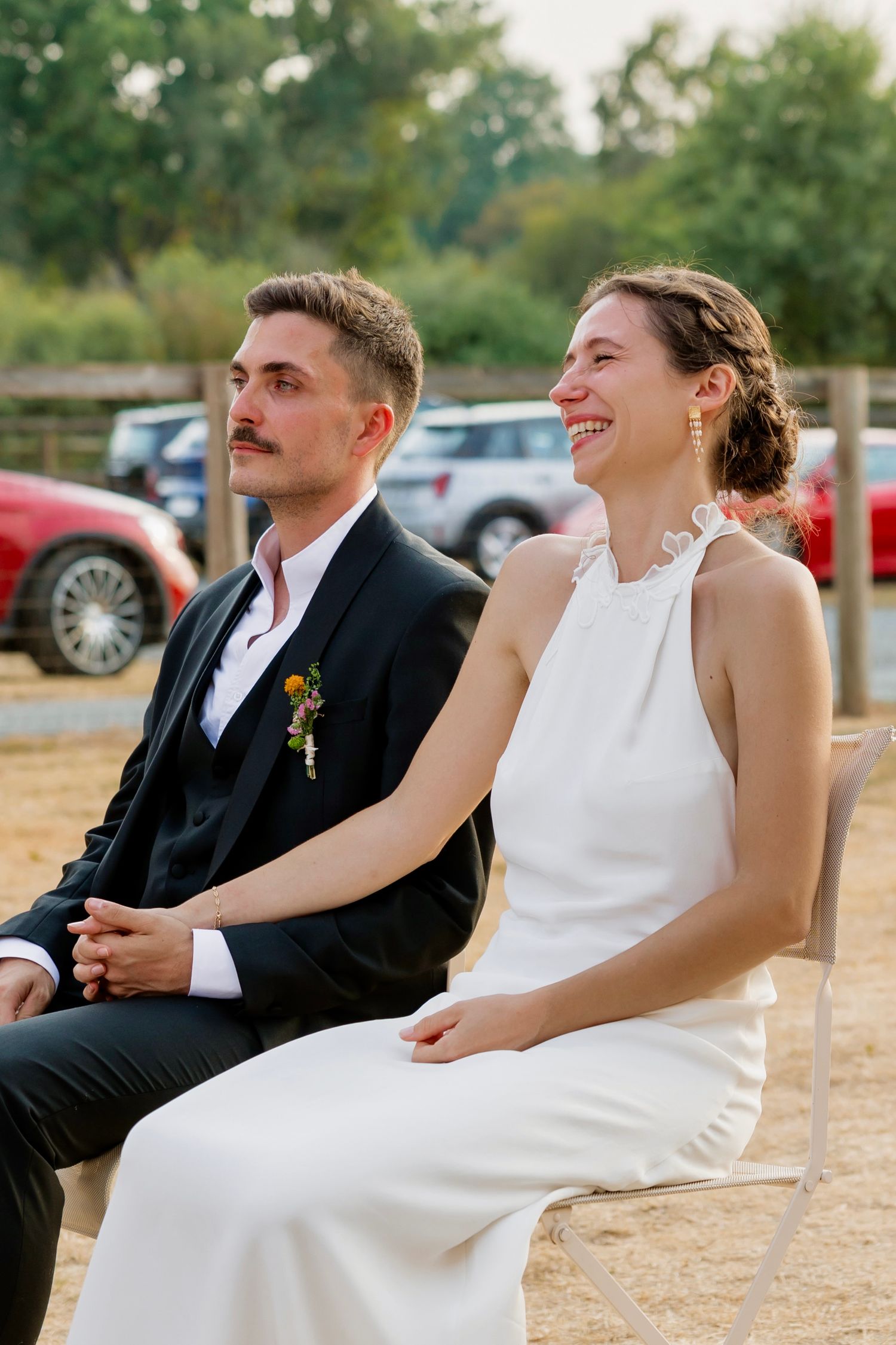 A smiling bride in white halter gown and groom in black suit with boutonniere seated outdoors at a wedding ceremony.
