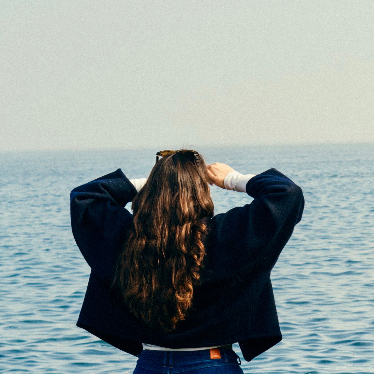 Personne aux longs cheveux bruns, face à la vue sur l’océan, portant une veste bleu marine.