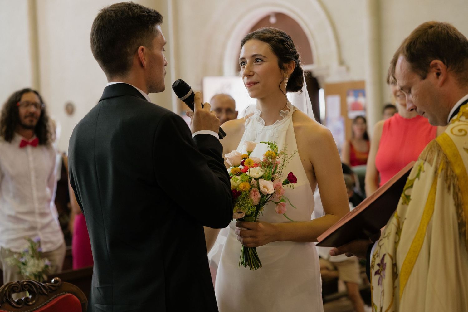 Bride holding colorful bouquet exchanges vows with groom at elegant church wedding ceremony.