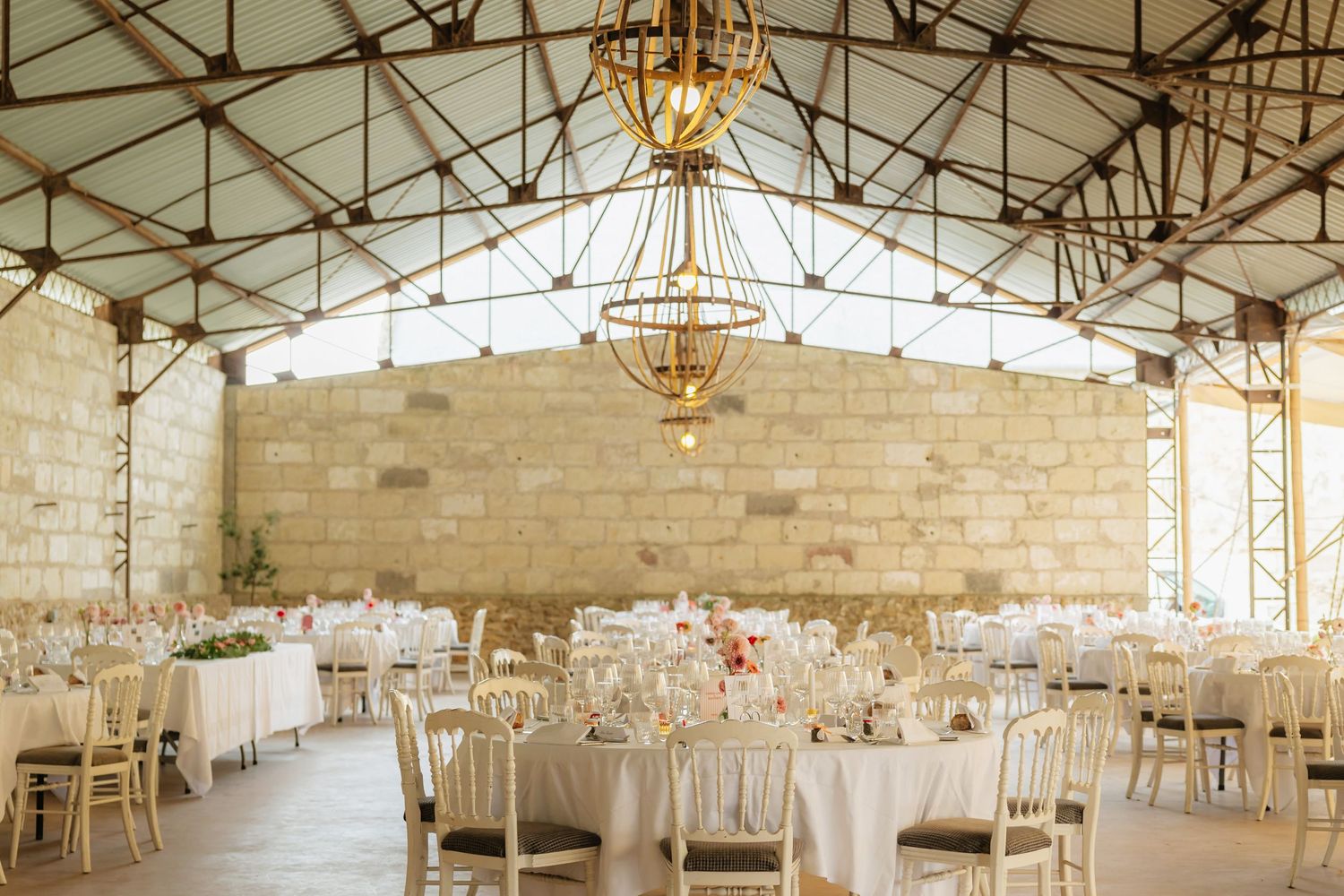 Elegant wedding reception hall with round tables, white linens, chandelier, and stone walls under a glass roof.