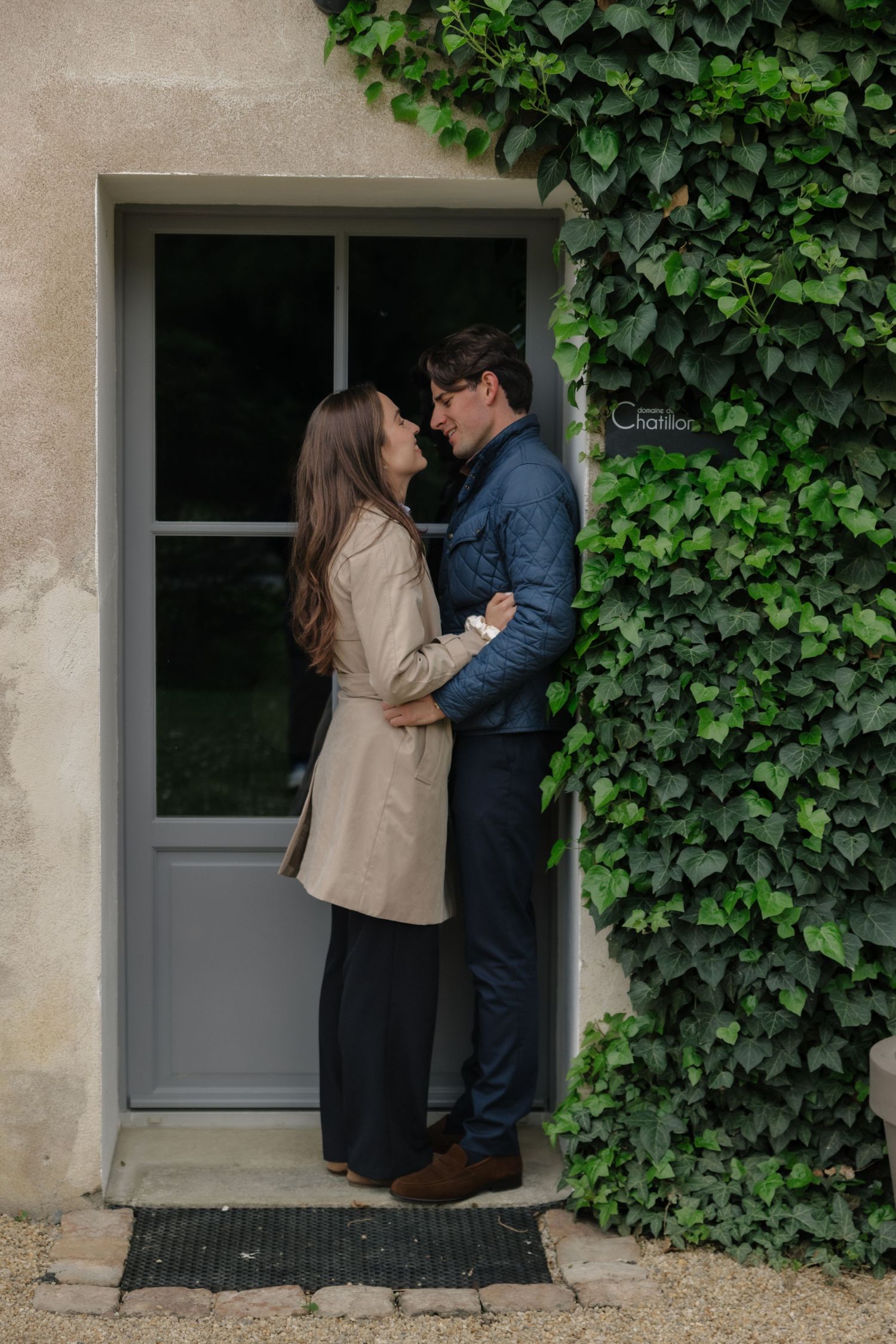Couple embracing romantically in a doorway covered with ivy, wearing casual elegant attire outdoors.