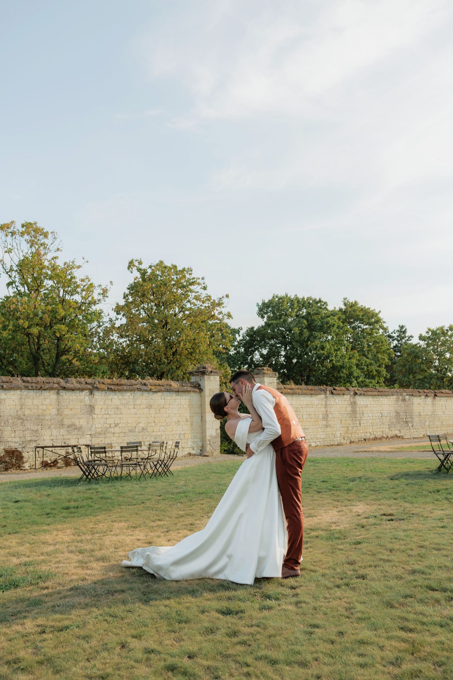 Groom in burgundy pants dips bride in white wedding gown for a romantic kiss in a lush garden setting.