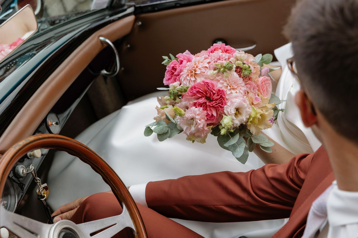A groom in a burgundy suit holds a pink floral bouquet while sitting in a vintage convertible car.