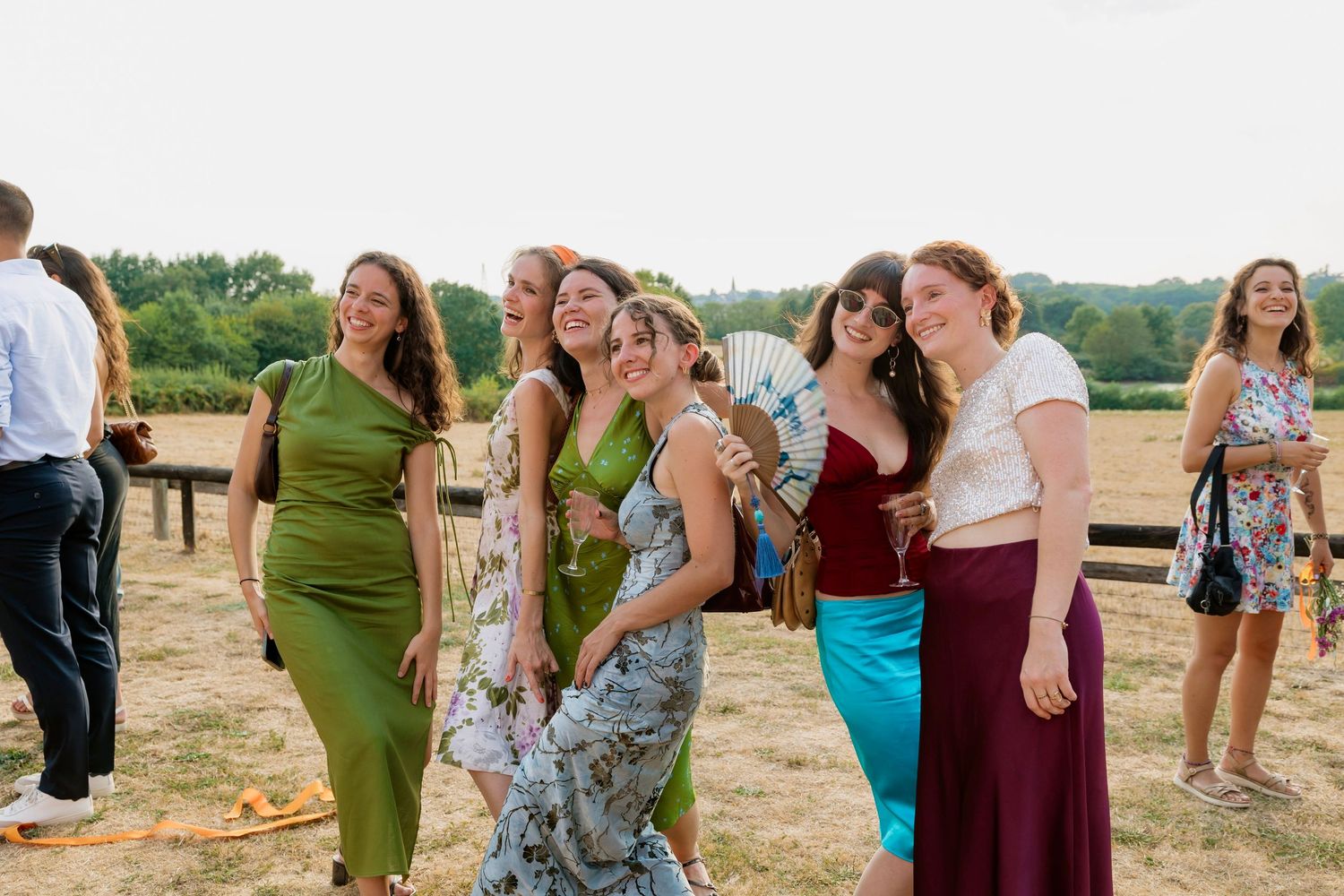 A group of women in colorful formal dresses laughing and posing together outdoors at a summer wedding reception.
