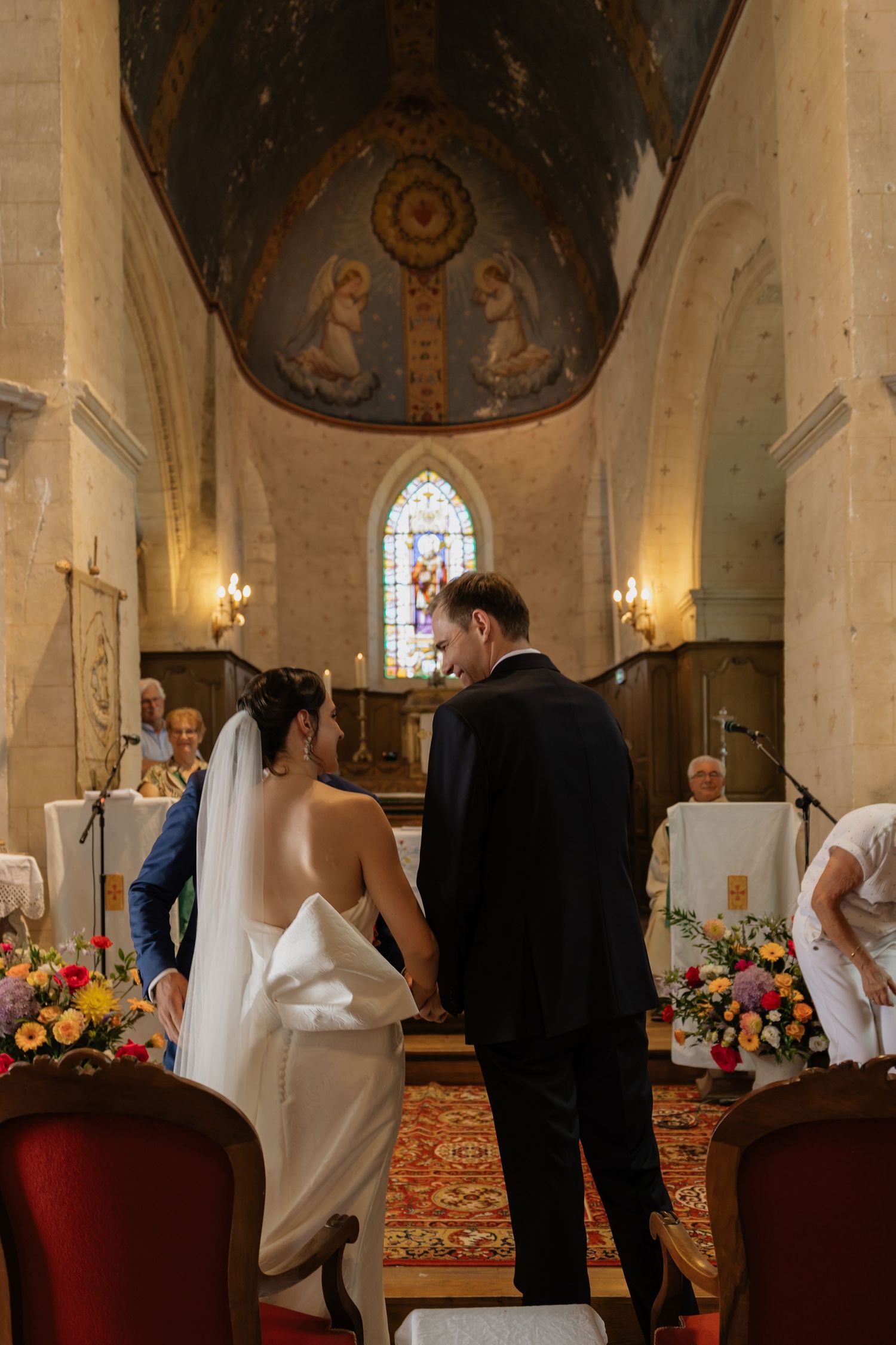 Bride and groom holding hands at church altar during wedding ceremony with colorful floral arrangements and Byzantine fresco.