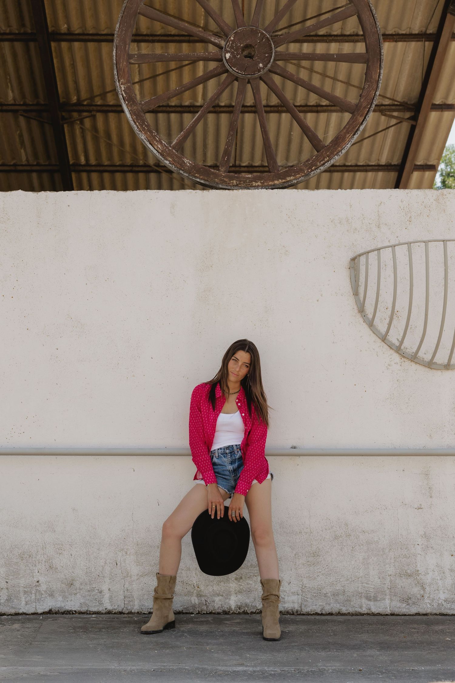 Young woman in red jacket, denim shorts, and tan boots holds black hat, posing against white wall with vintage wagon wheel above.