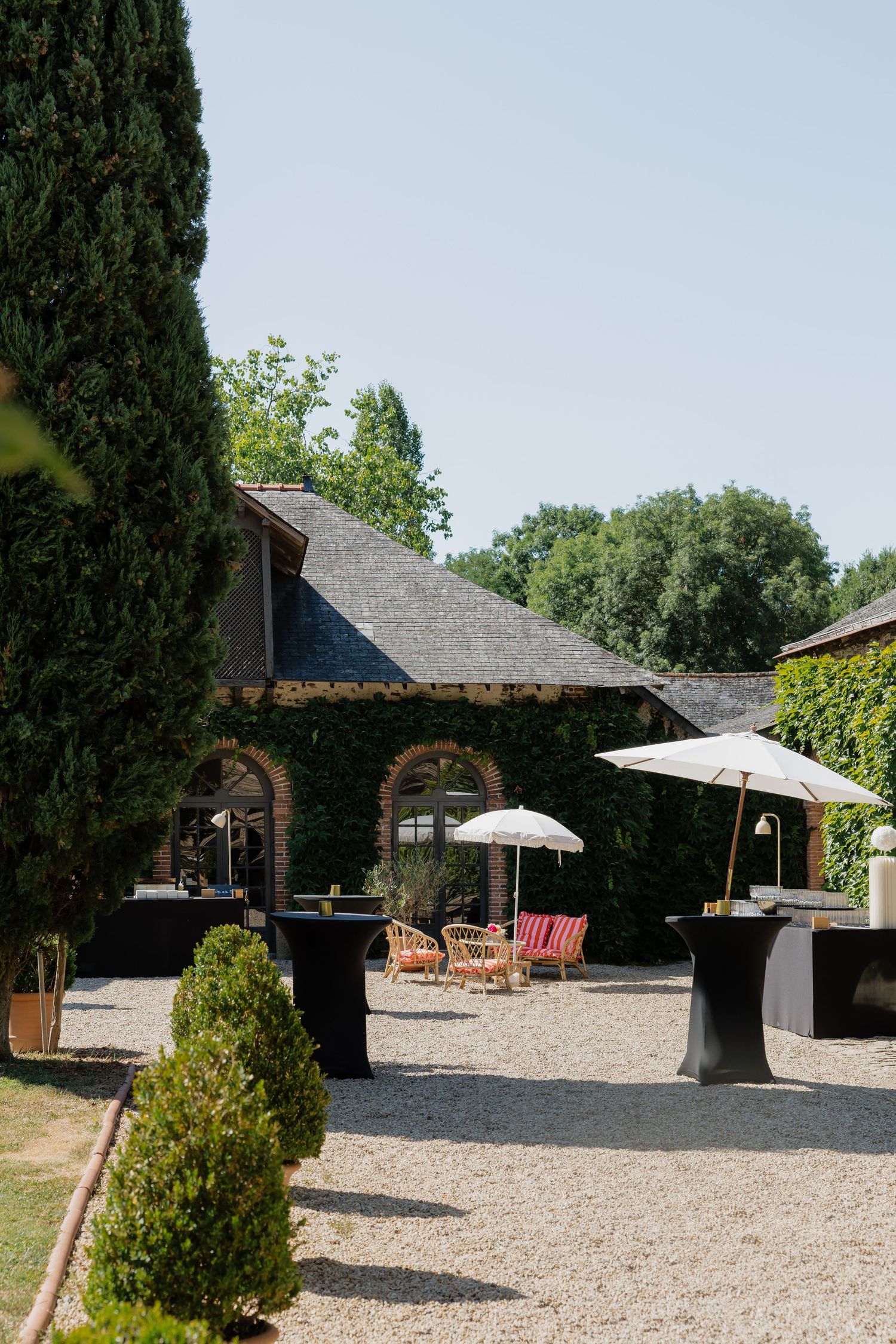 Elegant outdoor venue with black cocktail tables, white umbrellas, and rattan chairs on gravel beside a rustic brick building.