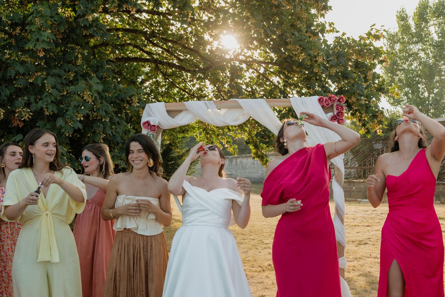 Bride tosses bouquet surrounded by bridesmaids in colorful dresses at outdoor golden hour wedding celebration.