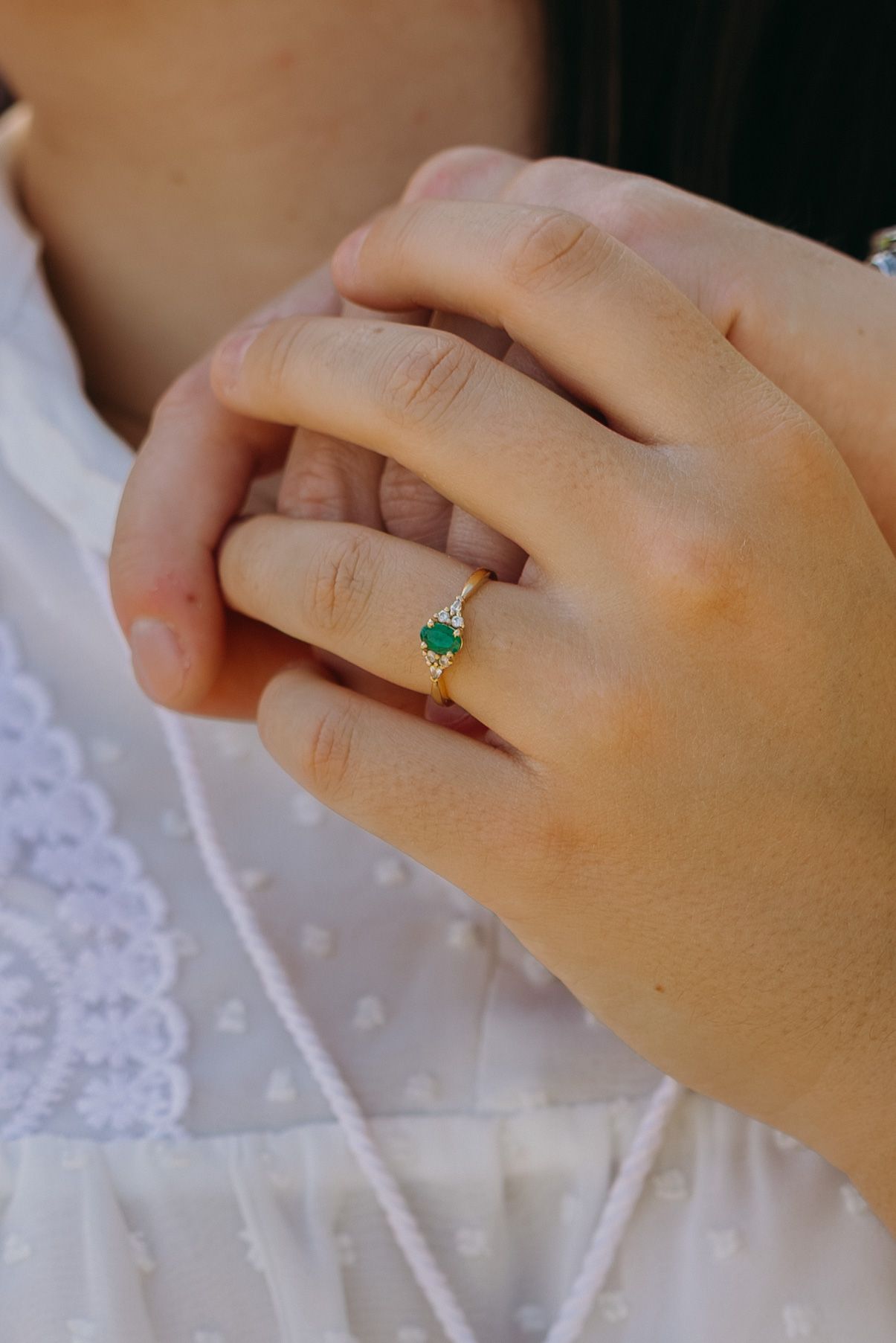 Close-up of hands showing an emerald engagement ring against white lace fabric.