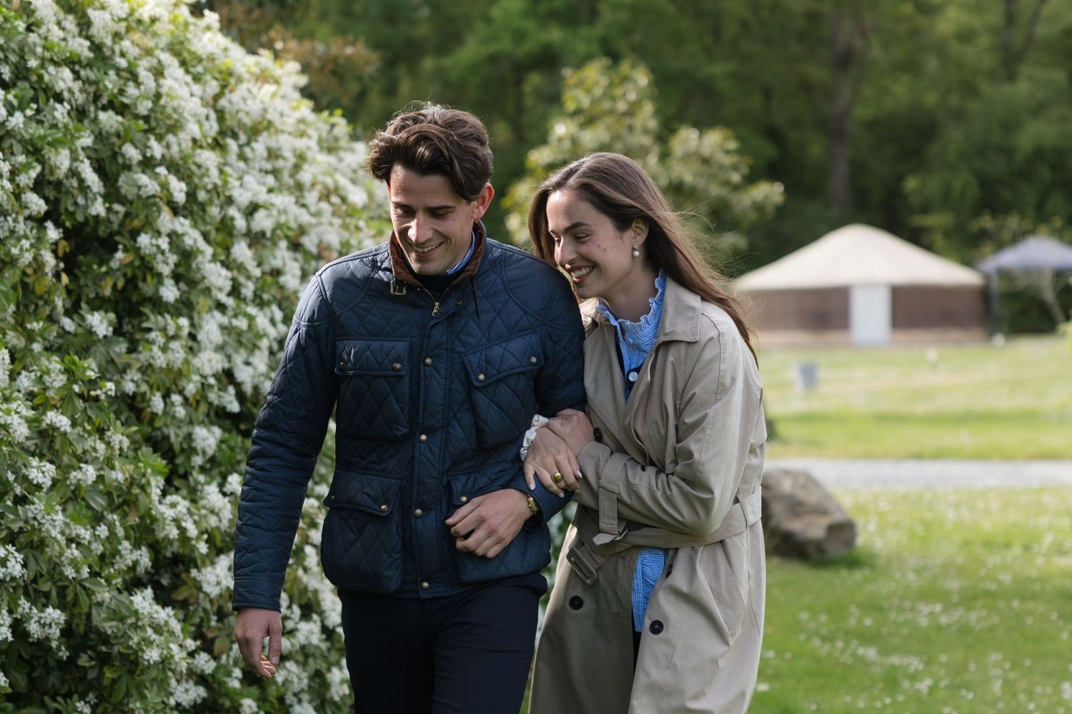 Couple walking arm in arm outdoors near blooming white flowers, smiling together on a green lawn.