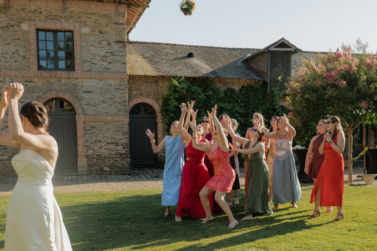 Bride tossing bouquet to excited guests in colorful dresses outside a rustic stone venue on a sunny day.