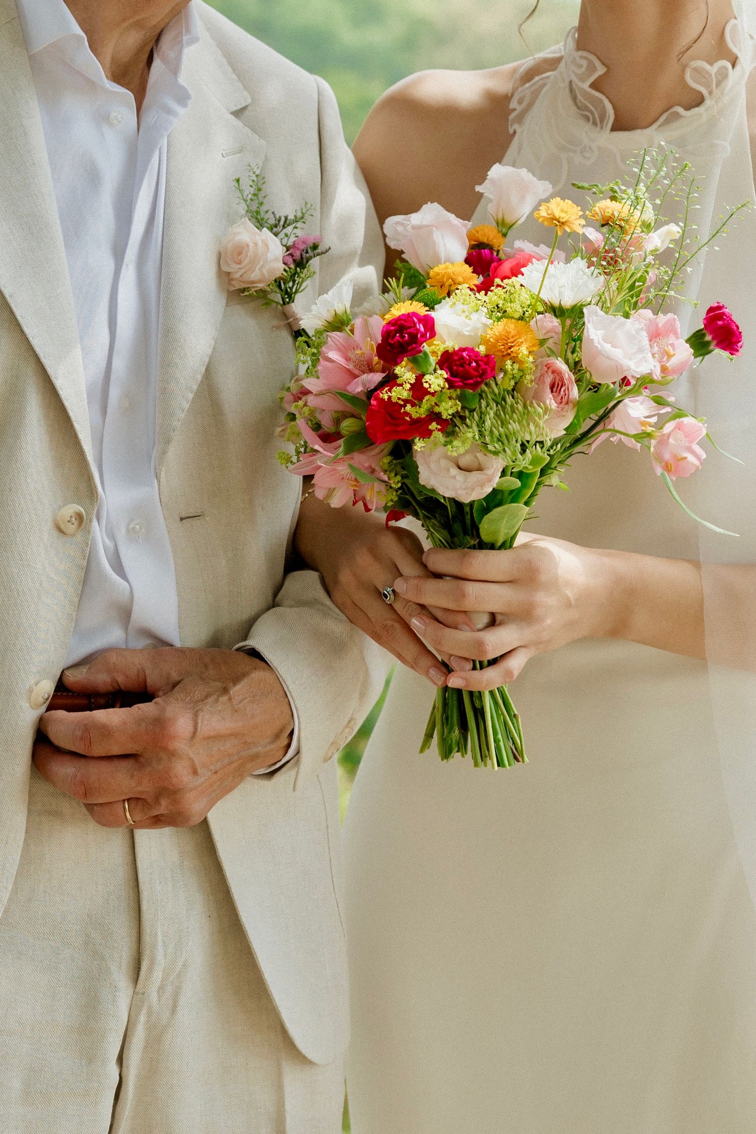 Bride holding colorful wedding bouquet with pink, red, and yellow flowers while standing beside groom in beige suit.
