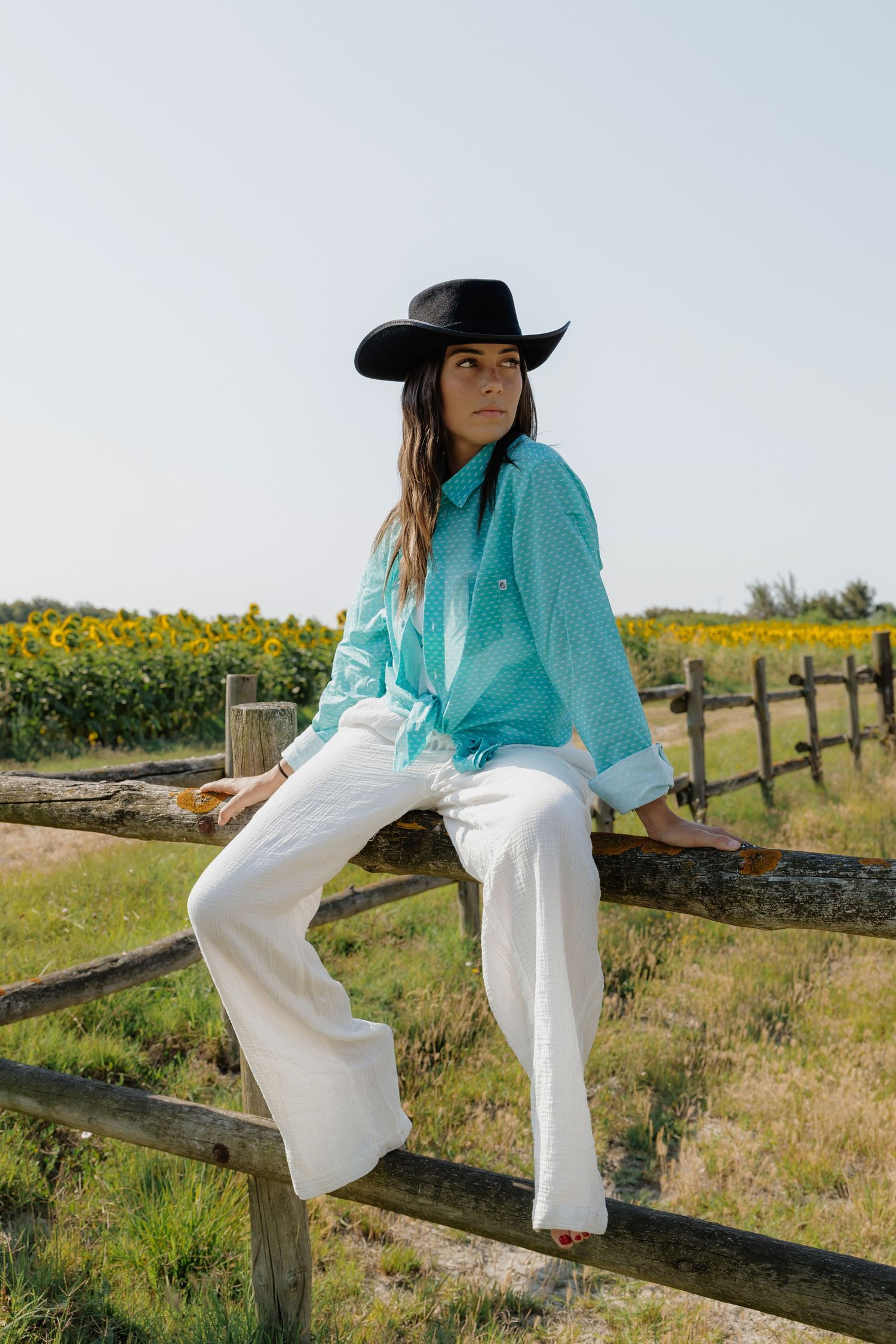 Woman in black cowboy hat, teal shirt, and white pants sitting on a wooden fence with sunflowers in background.