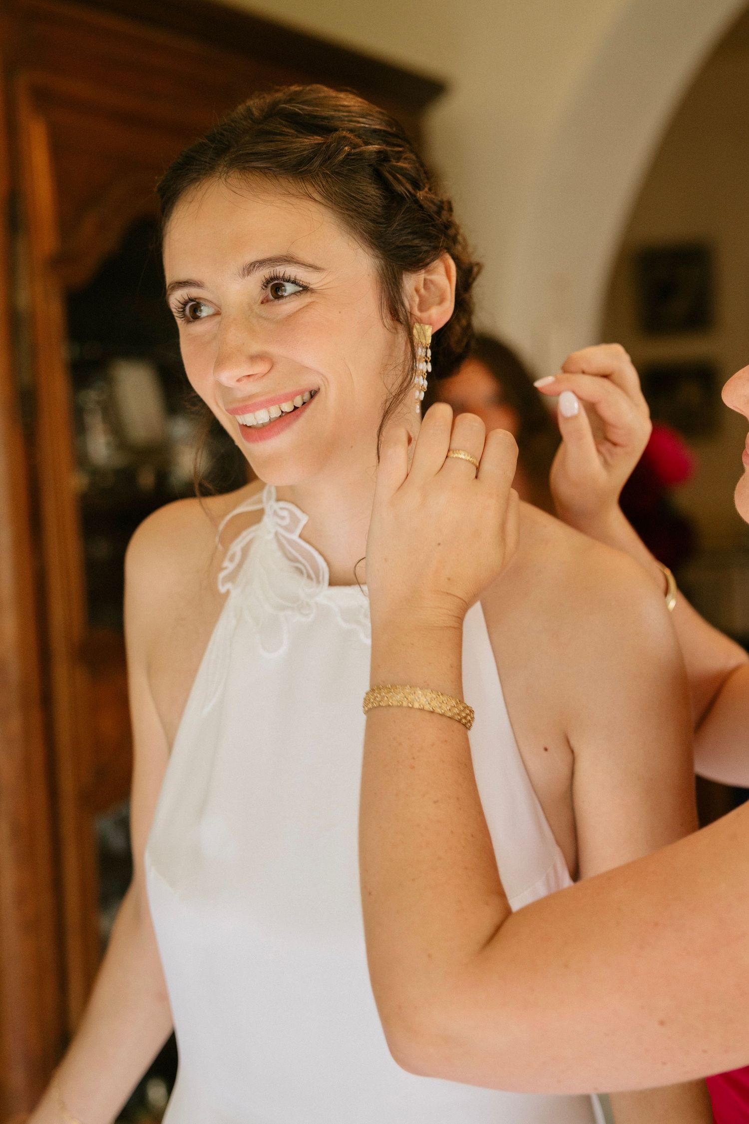 Smiling bride in white dress getting earrings put on, wearing gold bracelet and ring, preparing for wedding day.