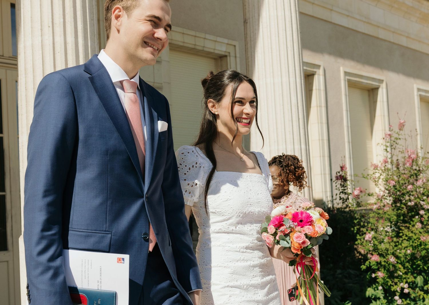 Smiling bride in white dress holding colorful bouquet stands with groom in navy suit outside elegant columned building.