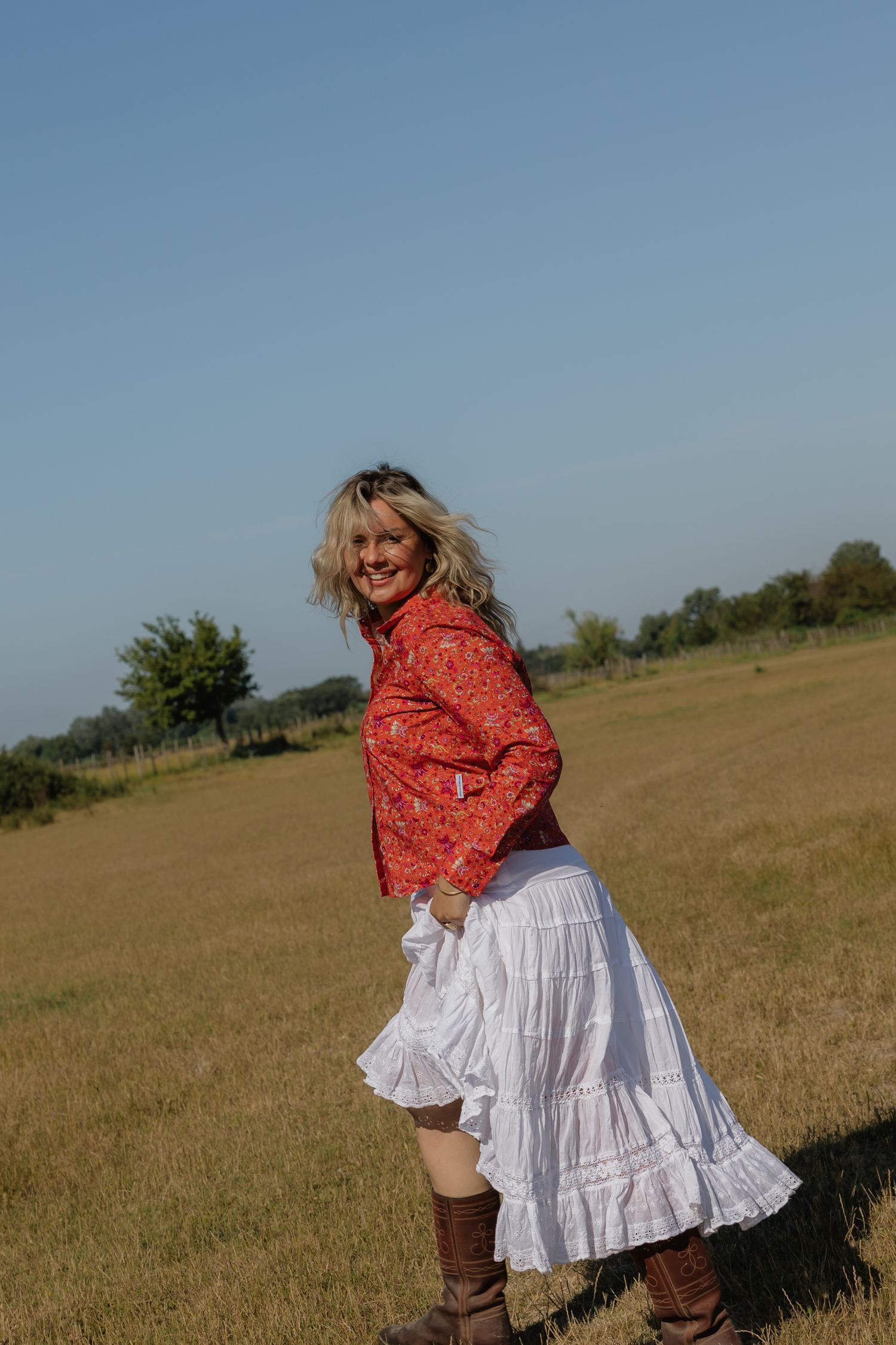 Smiling blonde woman in red floral top, white ruffled skirt, and brown boots posing in a sunny green field.