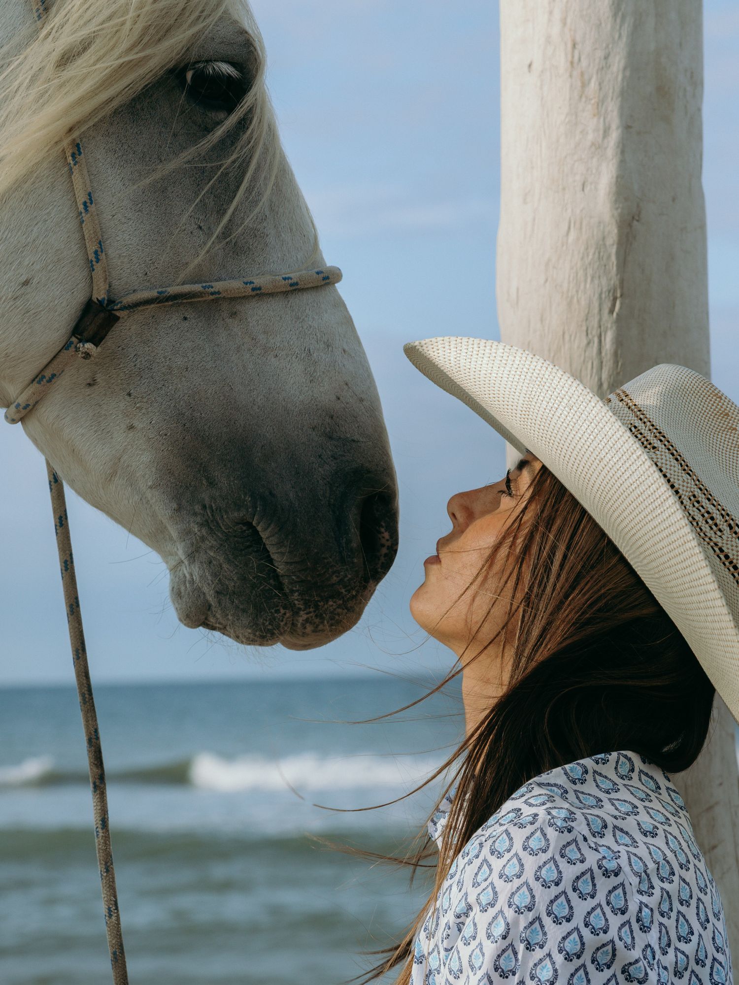 A person in a patterned dress and white hat shares an intimate moment with a white horse on a beach.
