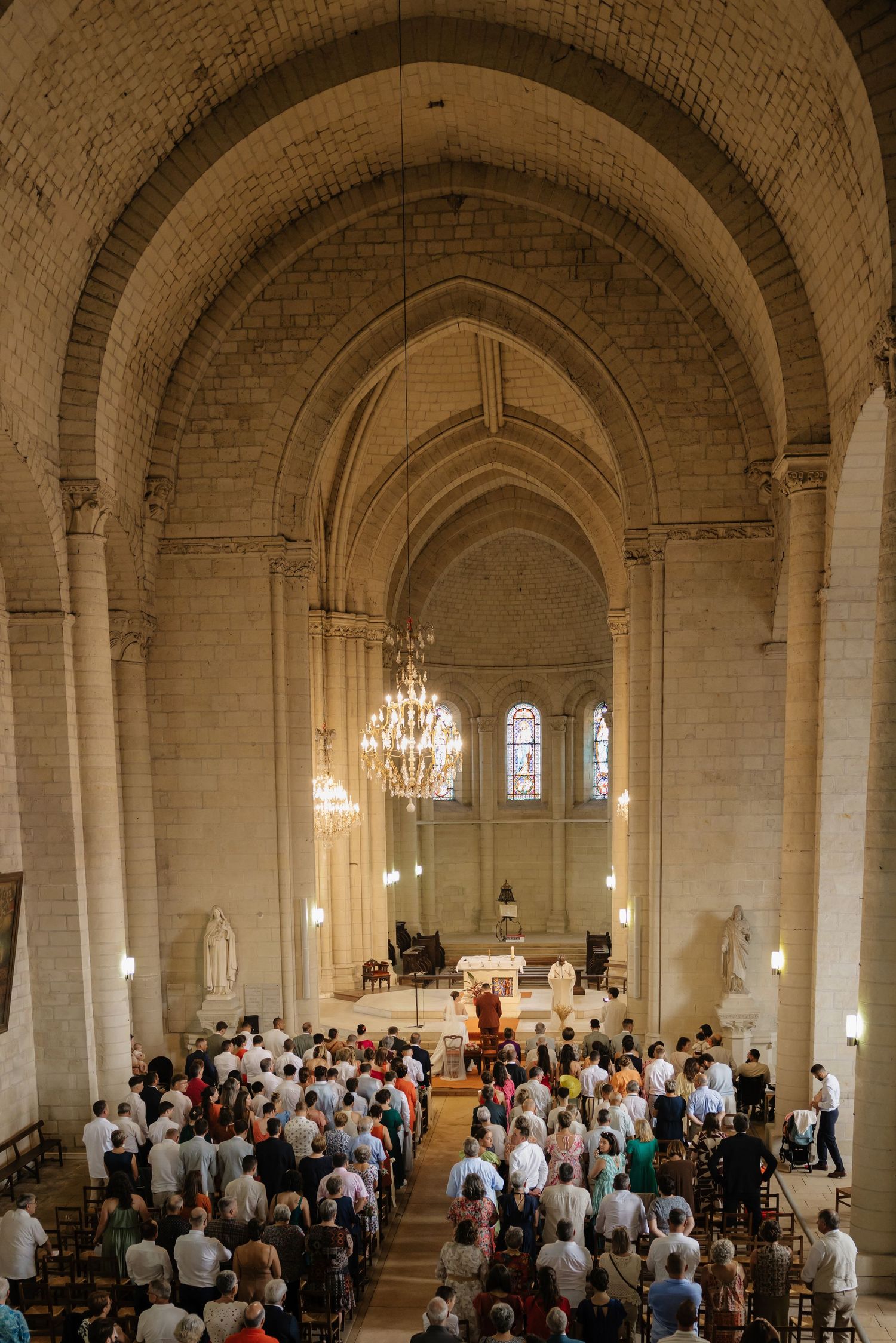 Congregation gathered inside a grand stone cathedral with arched ceilings and chandelier during a religious ceremony.