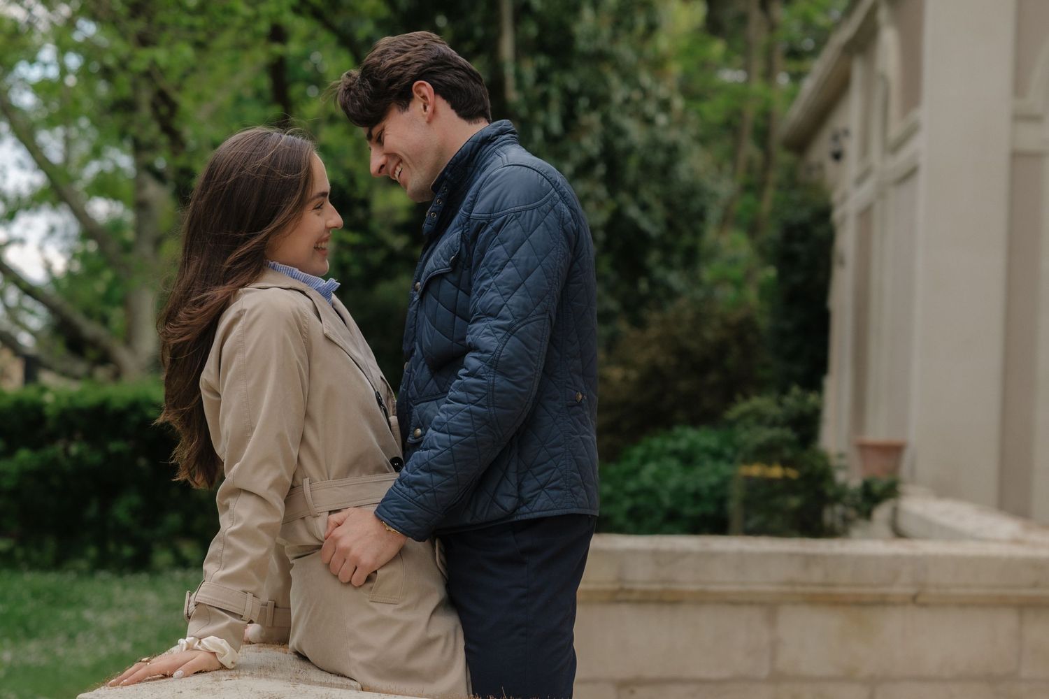A couple holds hands and gazes at each other outdoors, wearing casual jackets near a stone wall with greenery.