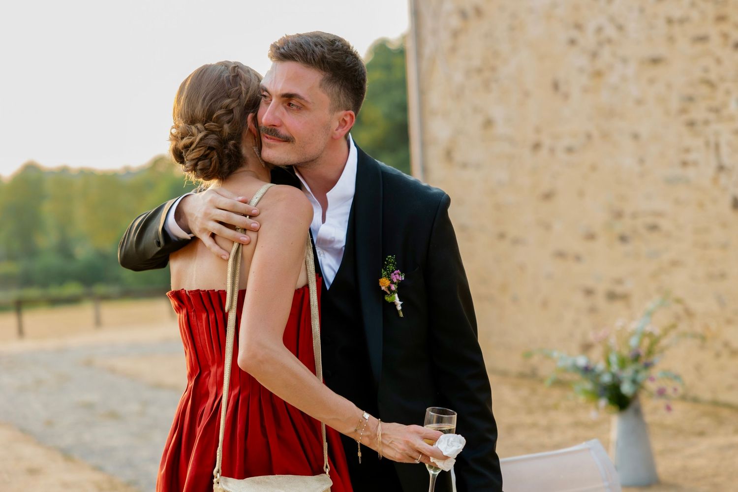 A young groom in a black suit kissing a woman in a red dress on the cheek at an outdoor wedding.