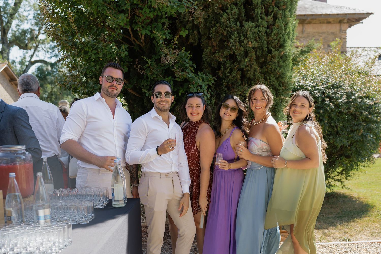Group of six friends smiling and posing outdoors at a summer wedding reception with greenery in the background.