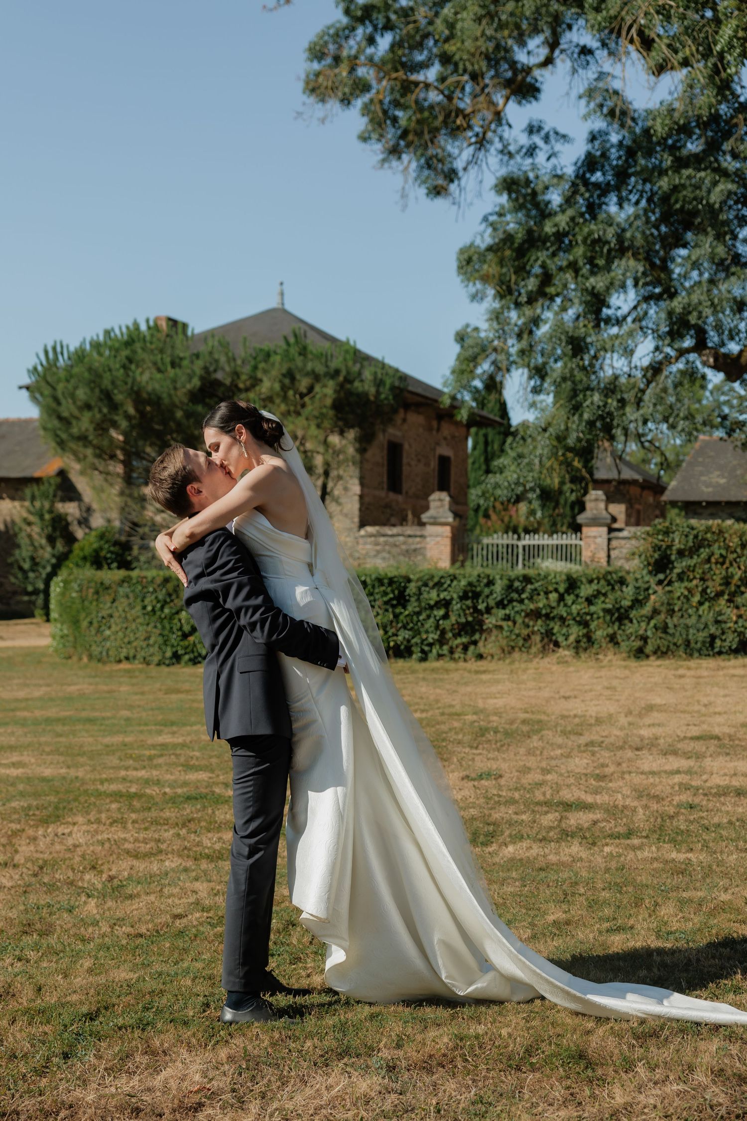 Groom lifts bride in white gown and veil on sunny garden lawn with lush greenery and chapel backdrop.