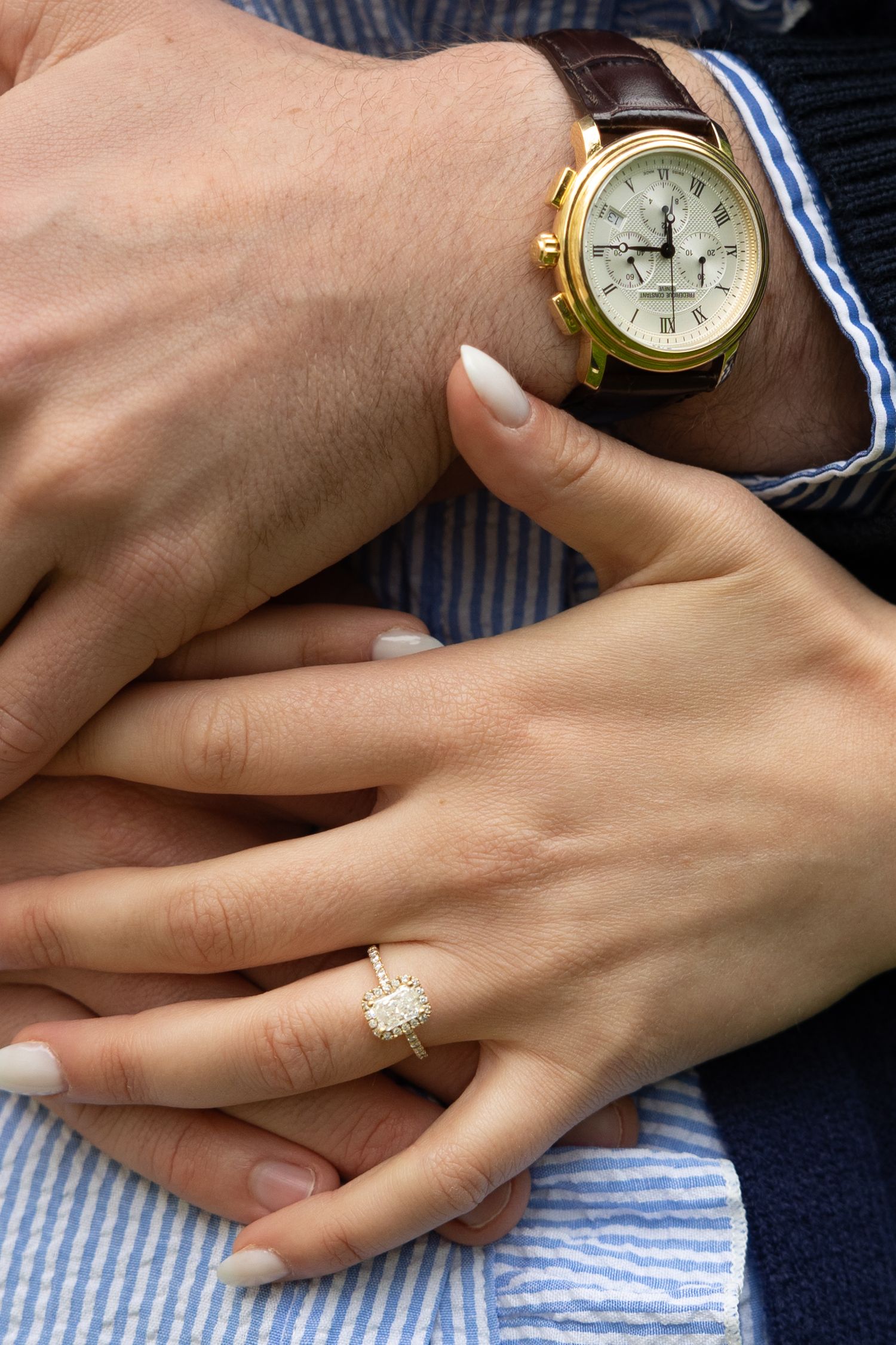 Close-up of couple's hands, woman wearing diamond engagement ring, man wearing gold chronograph watch with brown leather strap.