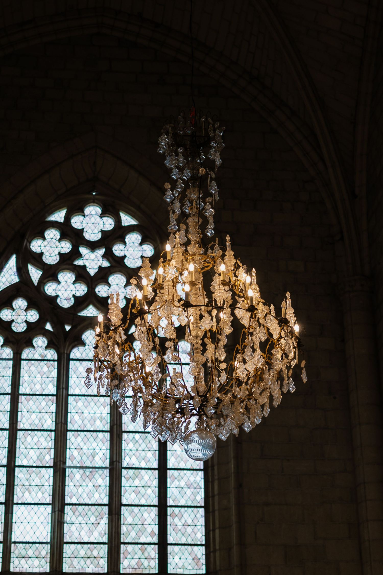 Ornate crystal chandelier hanging in a Gothic cathedral with arched stone ceiling and decorative stained glass windows.