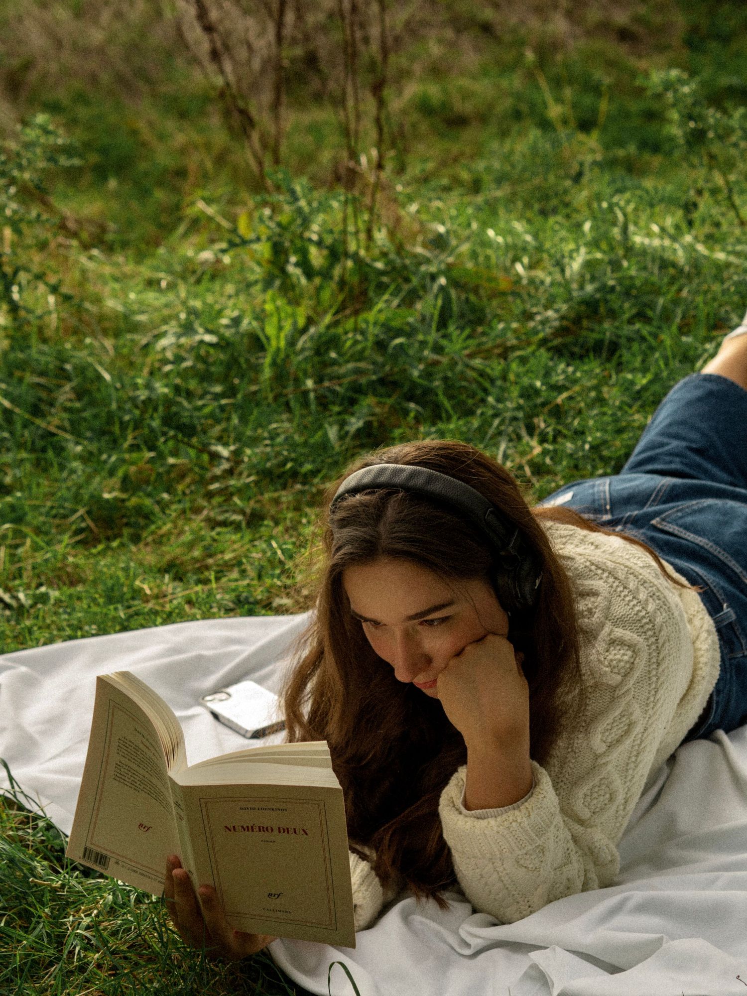 A person reads while lying on a white blanket in a grassy field.