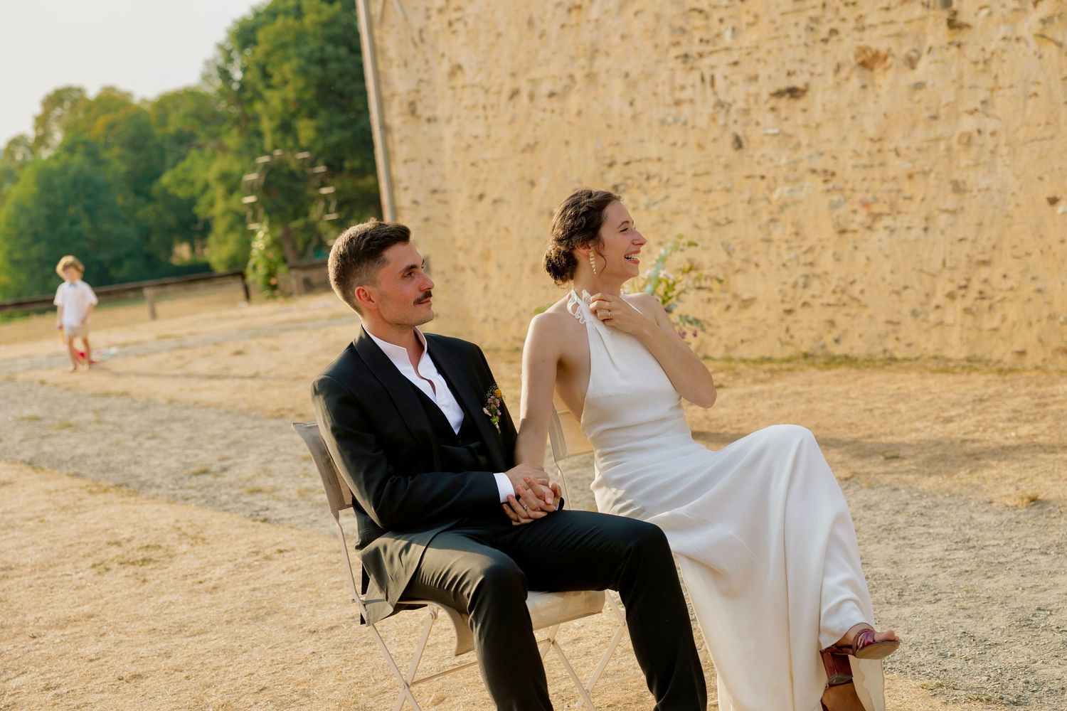 A bride and groom sit together on chairs outdoors near a stone wall, smiling joyfully on their wedding day.